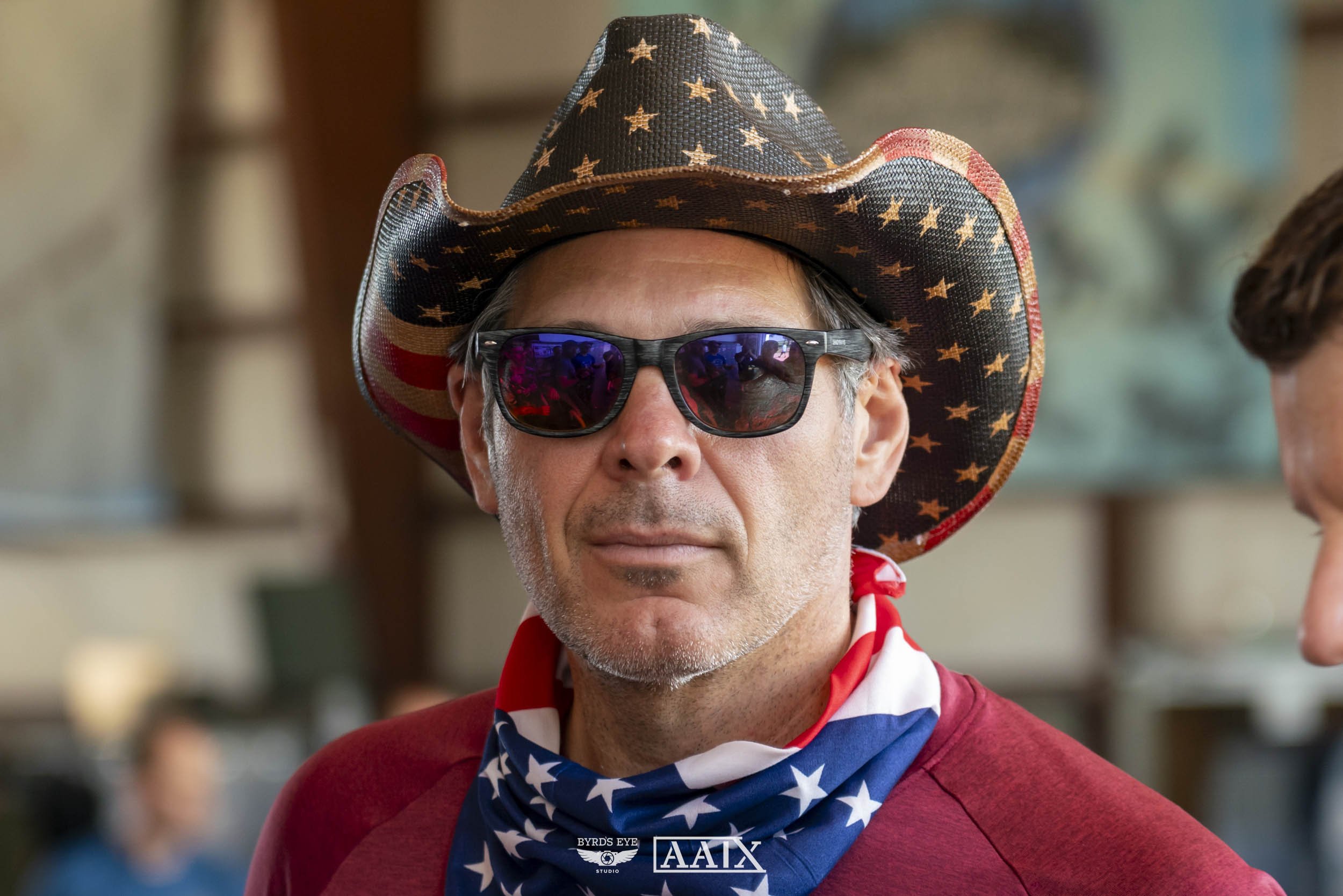 A middle-aged man with gray facial hair, wearing a patriotic cowboy hat decorated with stars and stripes, black sunglasses with reflective lenses, a red shirt, and an American flag bandana, at a gathering indoors.