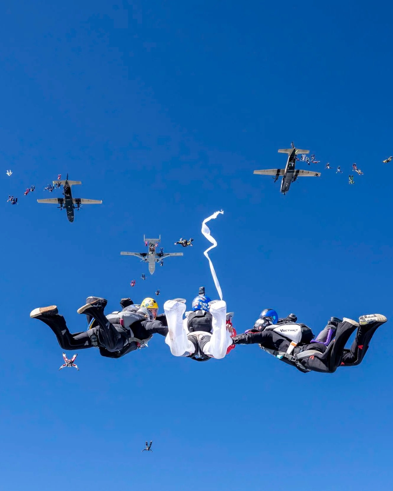 Three skydivers holding hands free-falling in a formation with airplanes and parachutes in the background.