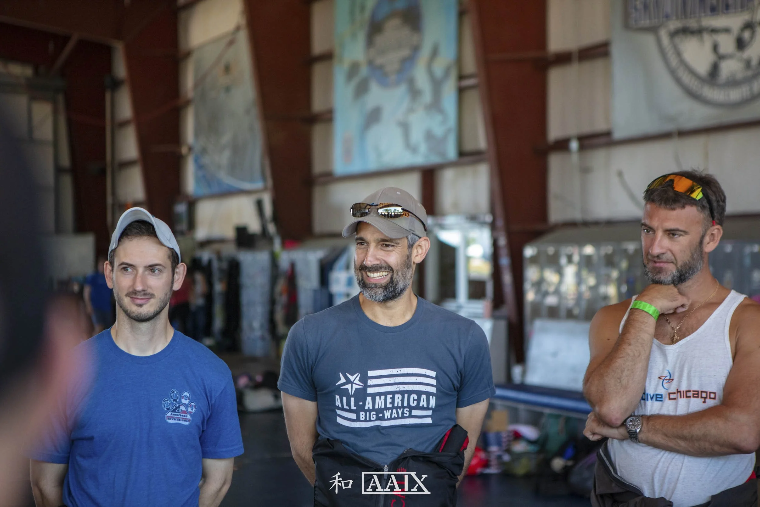 Three men standing indoors having a conversation, with a background that includes flags and maps. The man in the middle is wearing a gray baseball cap and a dark blue t-shirt, smiling. The man on the left is wearing a light blue cap and a blue t-shir