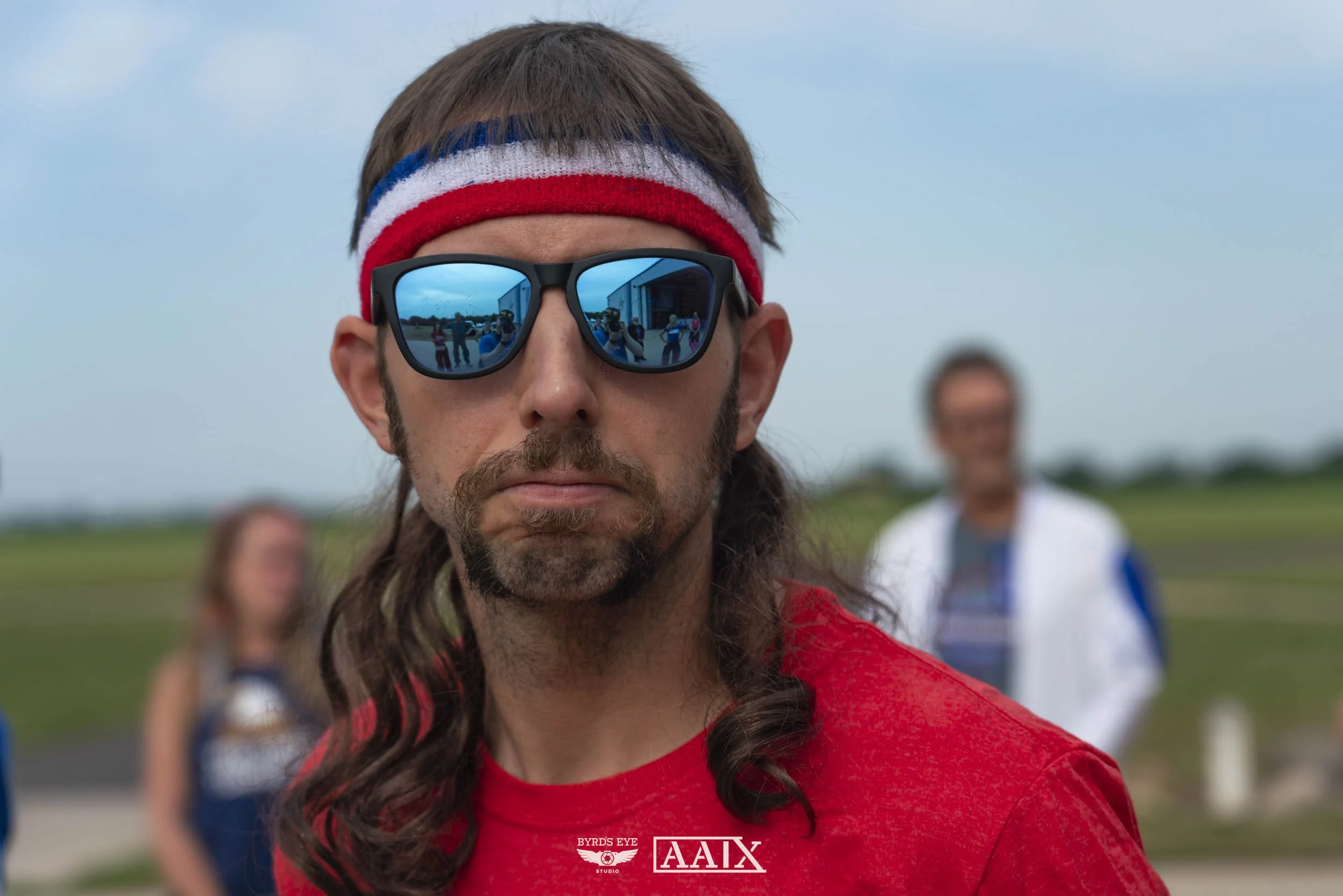 Close-up of a man with long hair, wearing reflective sunglasses and a red, white, and blue headband, standing outdoors with a field and people in the background.