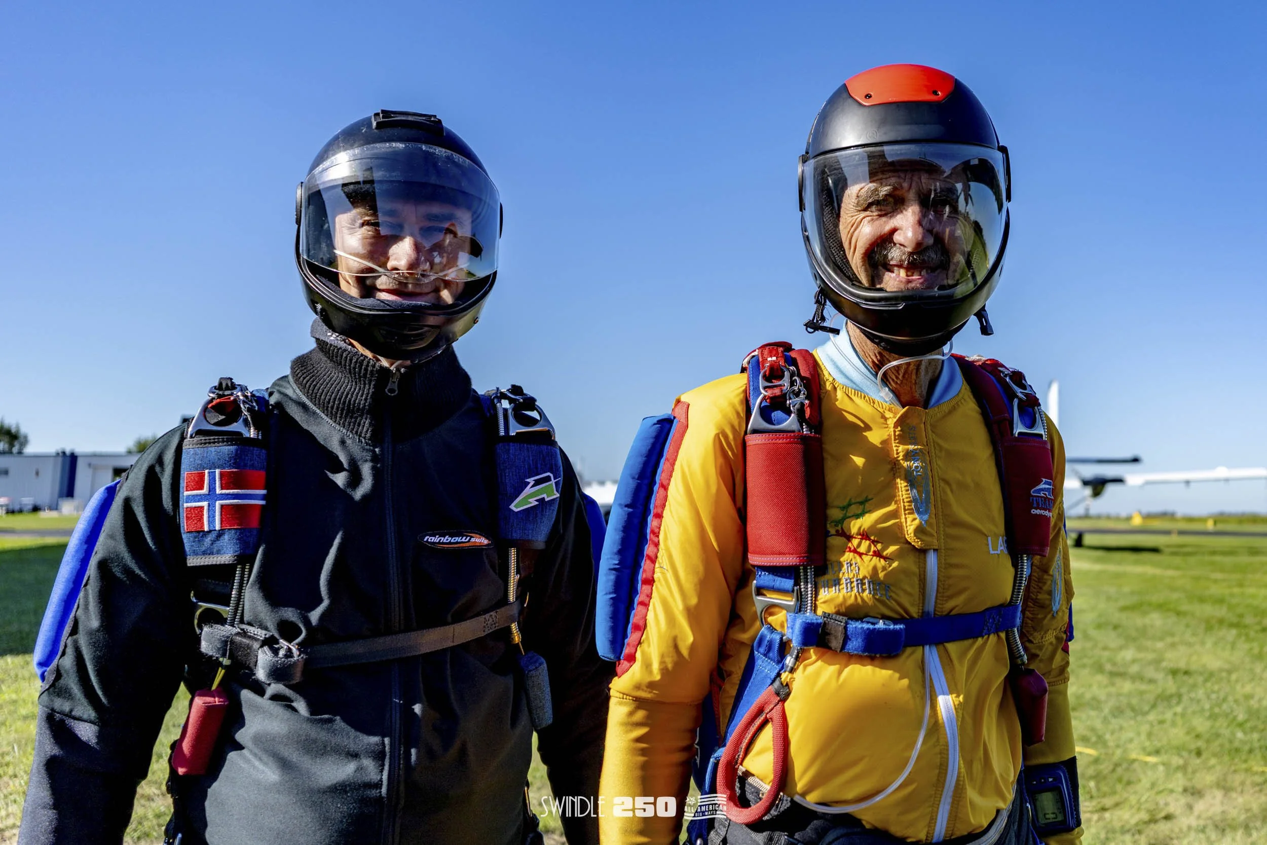 Two skydivers in helmets and jumpsuits smiling on a grassy field with a small airplane in the background under a clear blue sky.