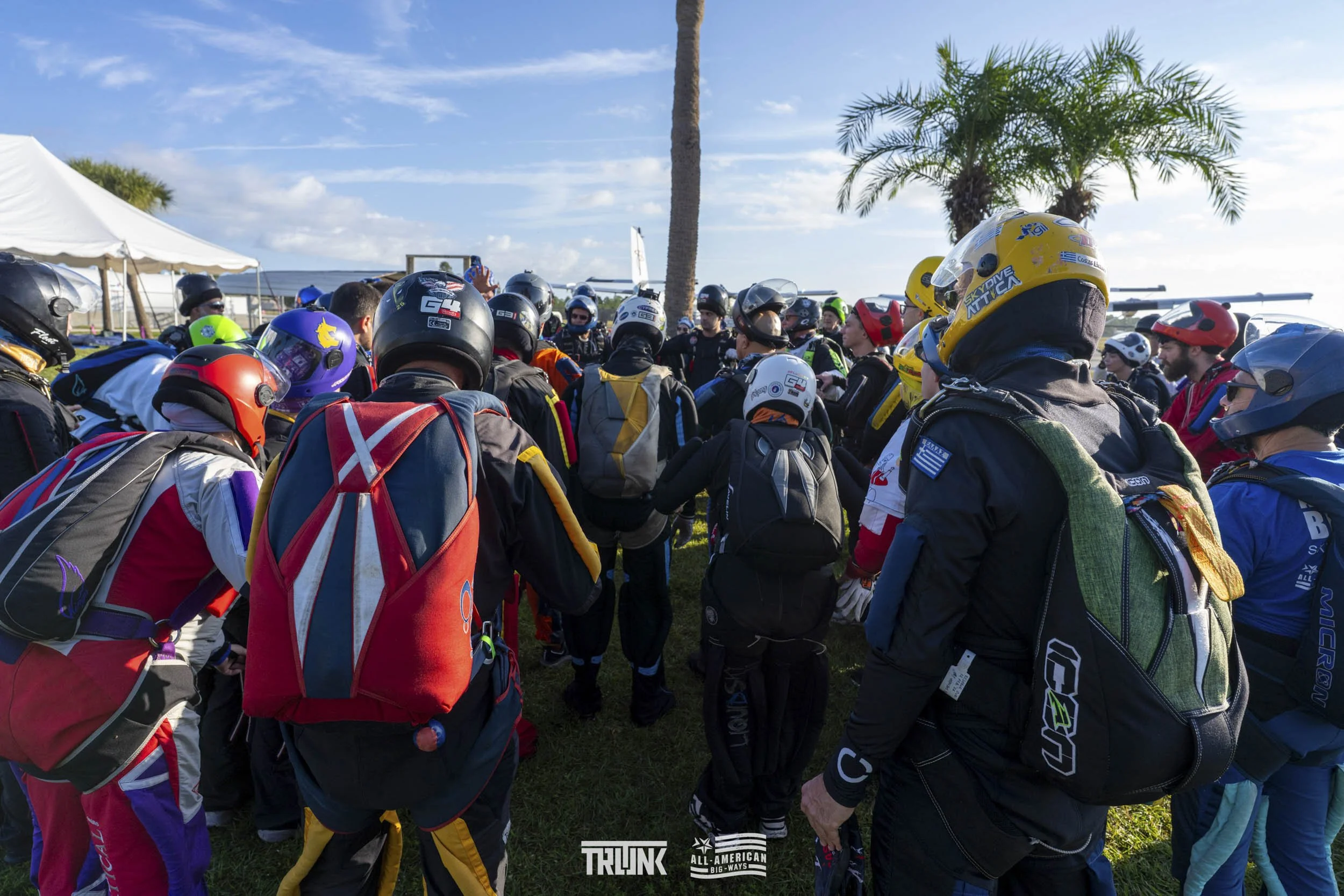 Group of skydivers gathered outdoors near palm trees, preparing for a jump on a sunny day.