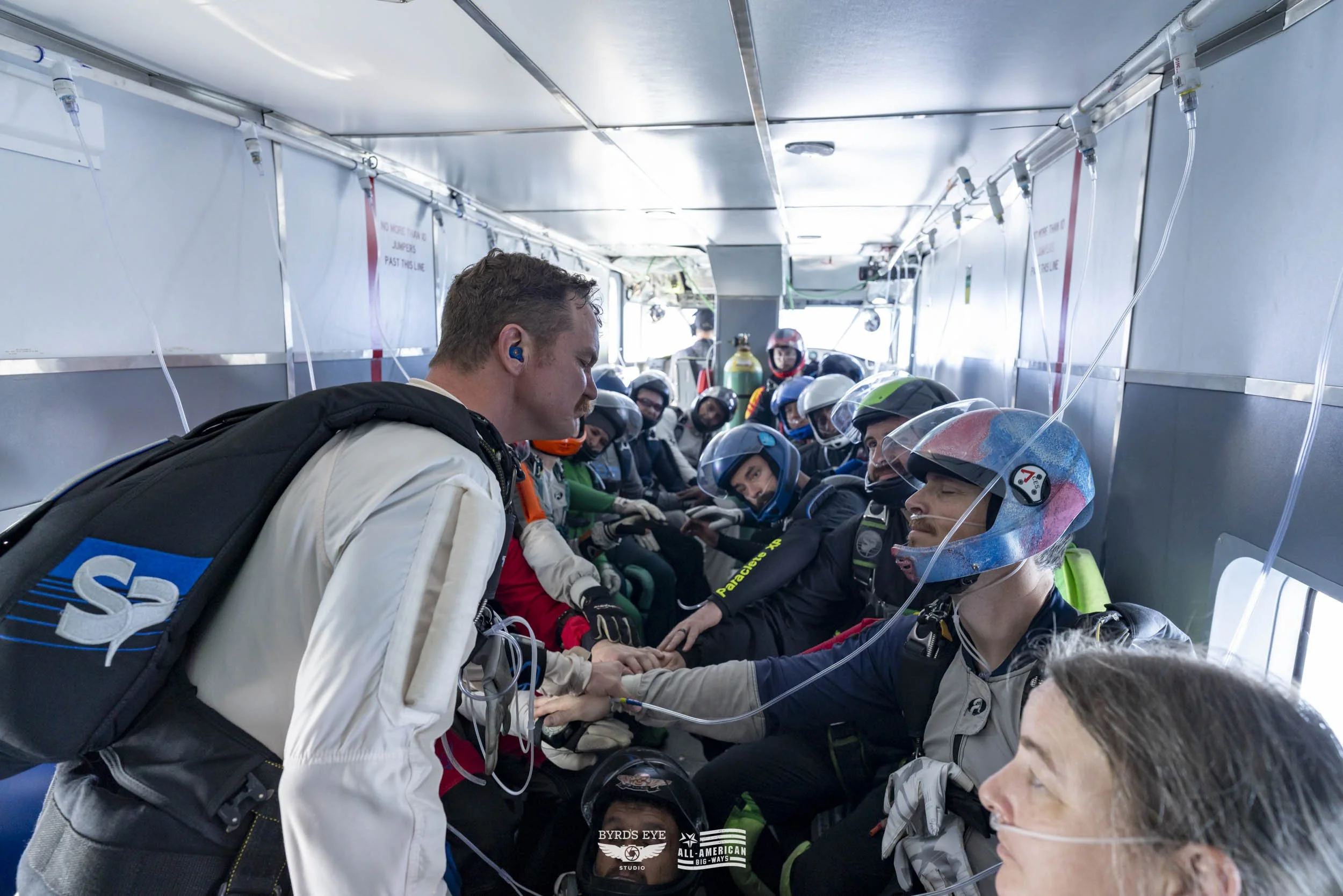 Skydivers seated inside the aircraft with a pilot, preparing for a jump, holding hands in a circle.