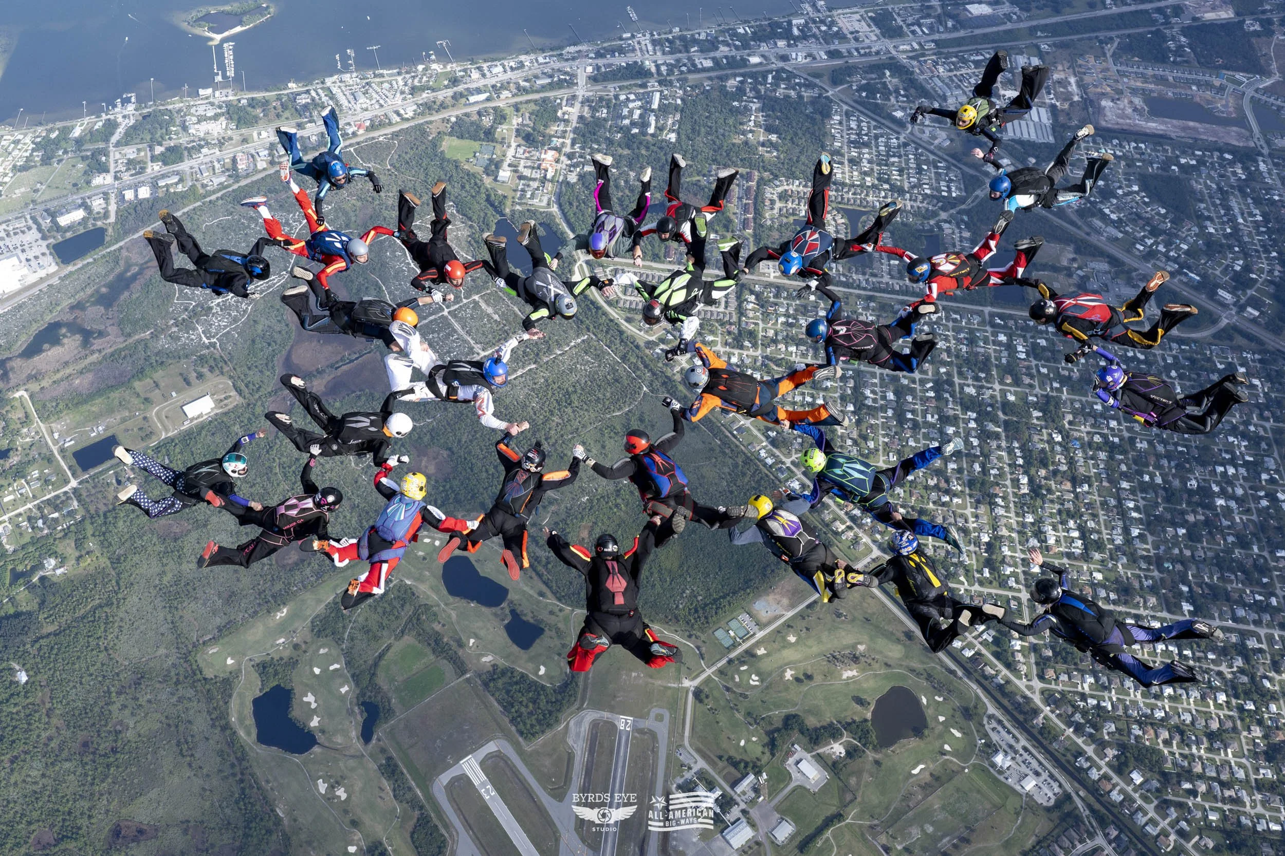 Group of skydivers holding hands in a circle during free fall above a city with parks, lakes, and buildings.