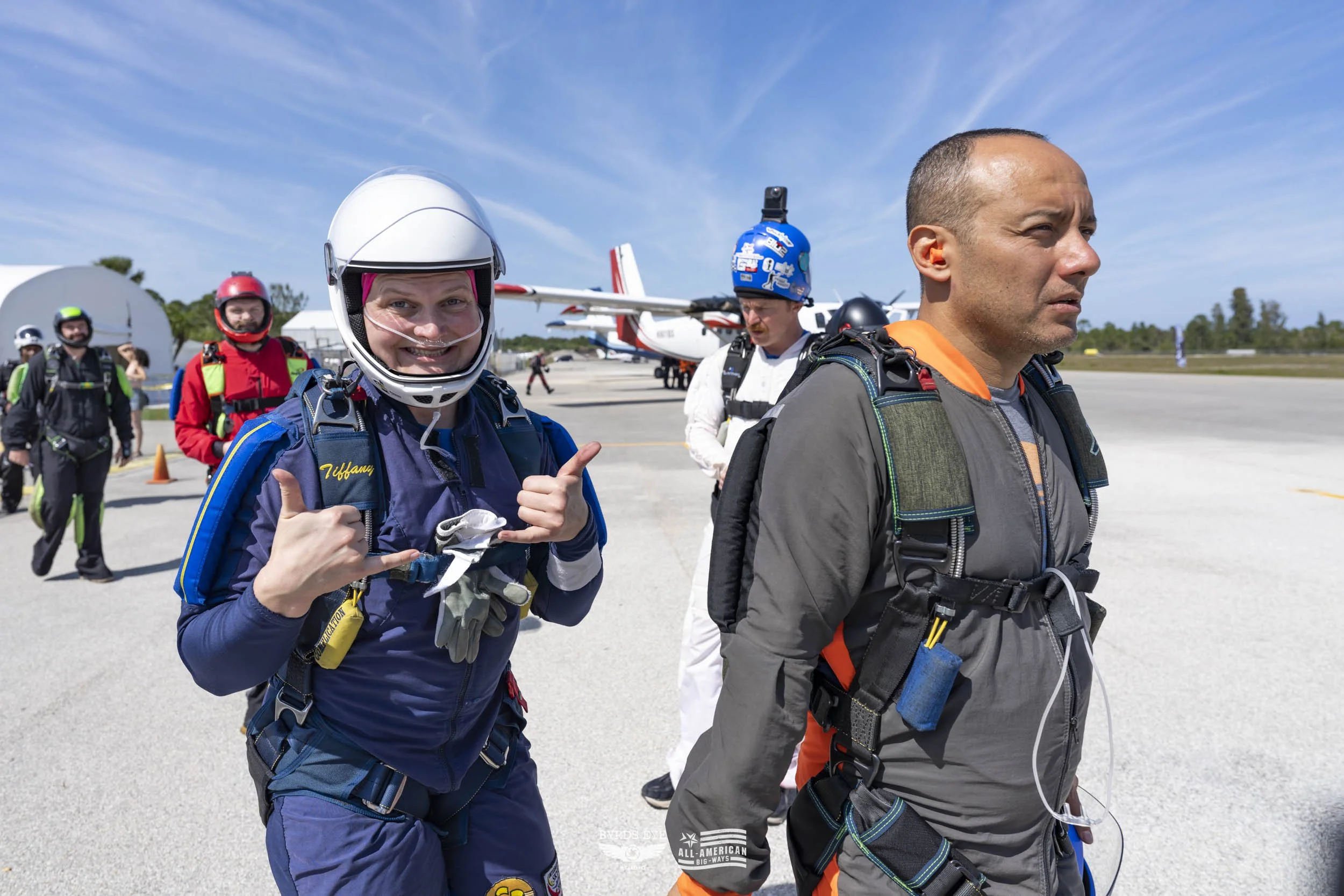 Skydivers on an airfield preparing for a jump, with a small aircraft in the background, clear sky, and people in jumpsuits and helmets.