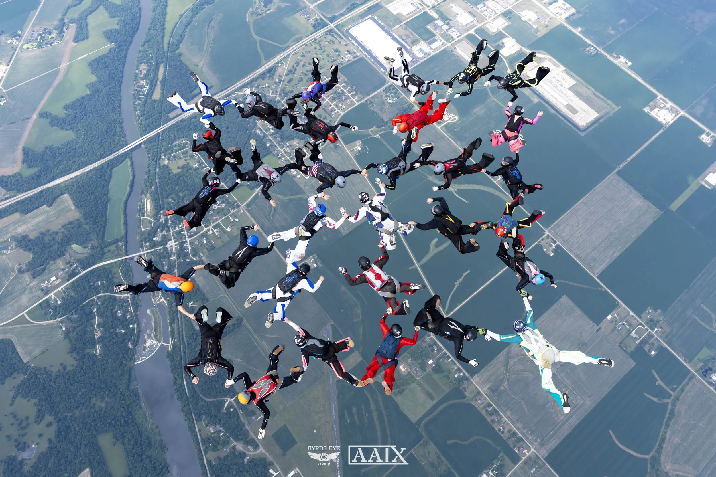 Group of skydivers in freefall formation over a landscape with water bodies, roads, and fields.
