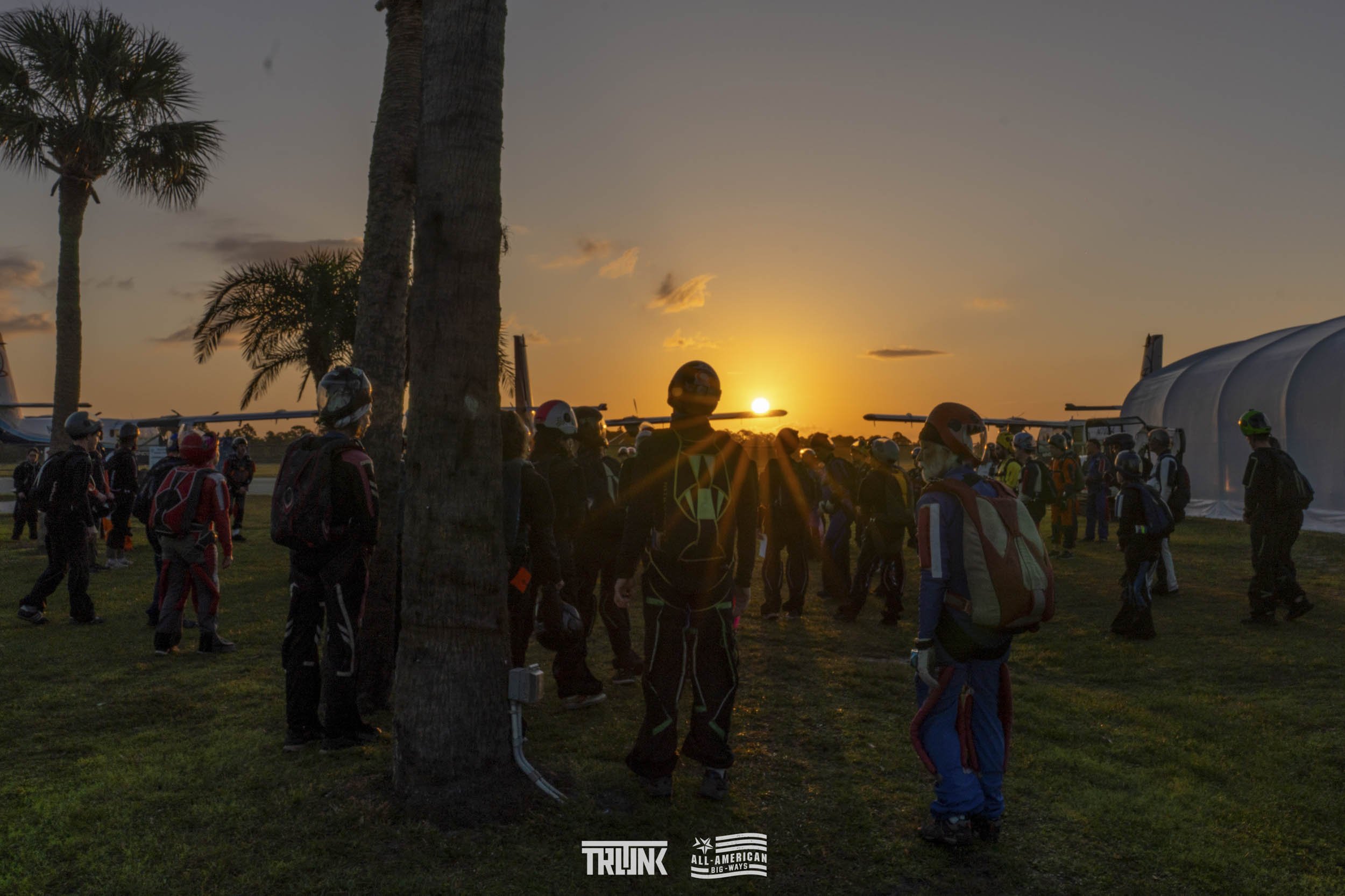 People dressed in skydiving gear gathering on a grassy area at sunset, with palm trees and a white tent in the background.