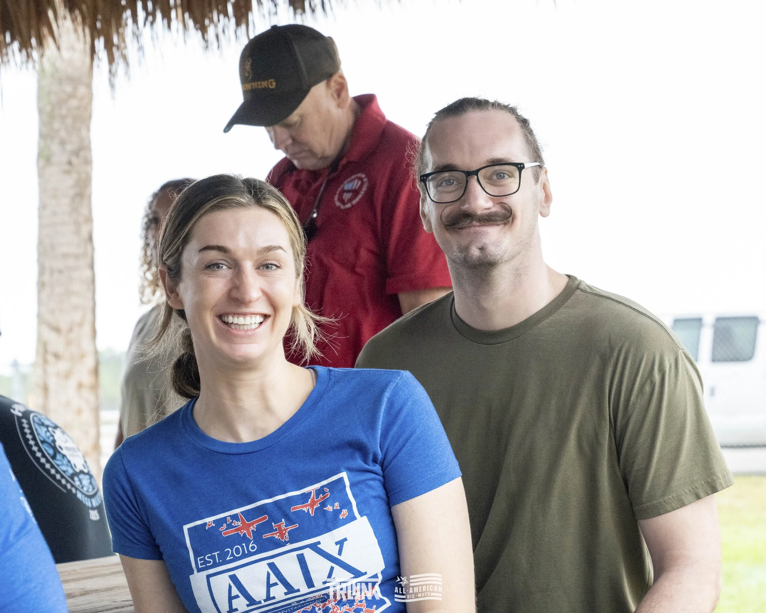 A group of smiling people at an outdoor event, with a woman wearing a blue t-shirt and a man in an olive green t-shirt in the foreground. In the background, a man in a red shirt and black cap is looking down.