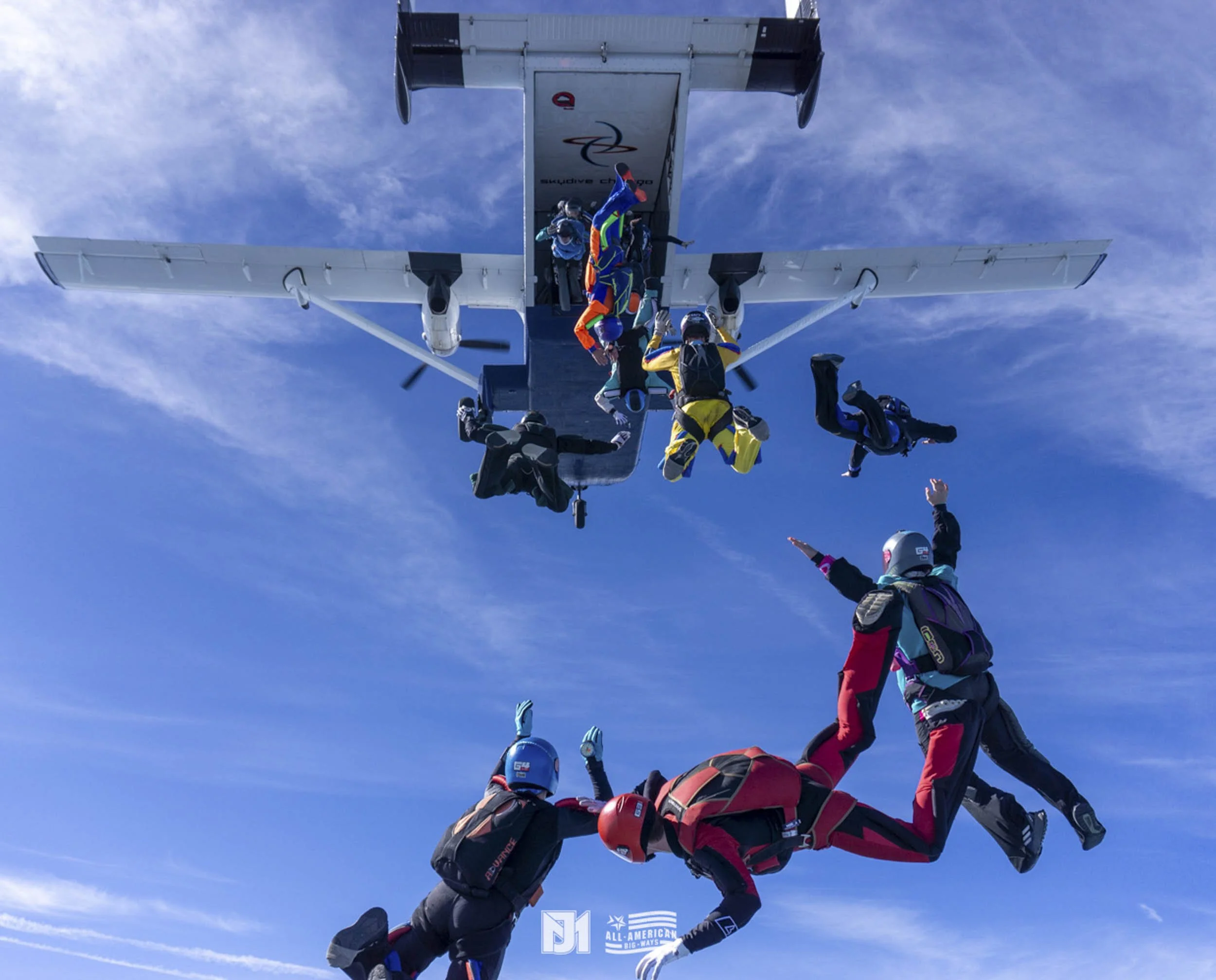 Skydivers in freefall after jumping from an airplane, with the plane visible above and a blue sky with wispy clouds in the background.