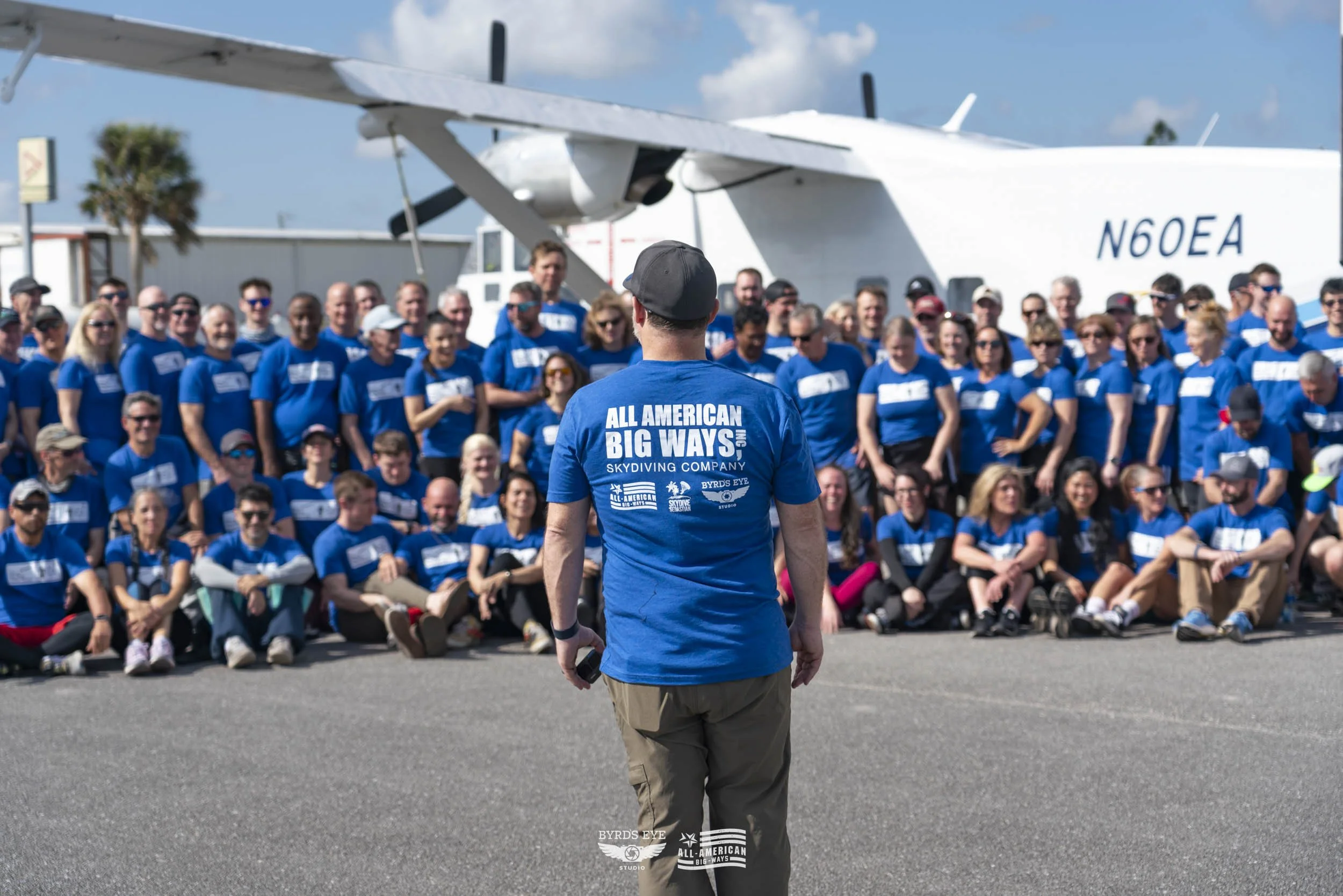 Group of people in blue shirts posing for a photo in front of a large airplane with the tail number N60EA. A person in the foreground wearing a blue shirt with the words "All American Big Ways Skydiving Company" on the back, holding a phone.