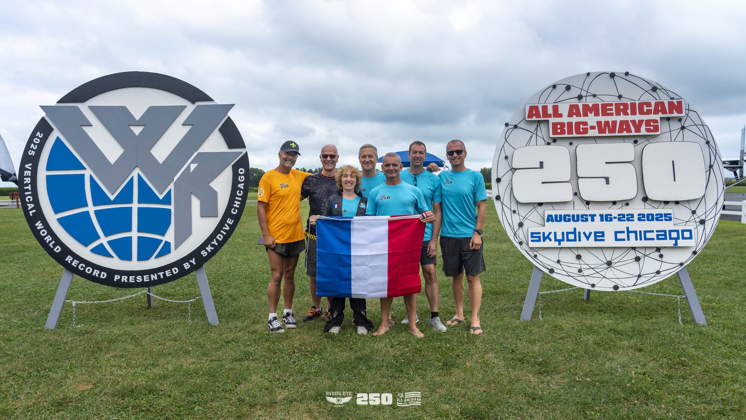 Group of seven skydivers standing on grass, holding a French flag, with large event signs in the background. The signs indicate a record for 250 skydivers at Skydive Chicago from August 16-22, 2023.