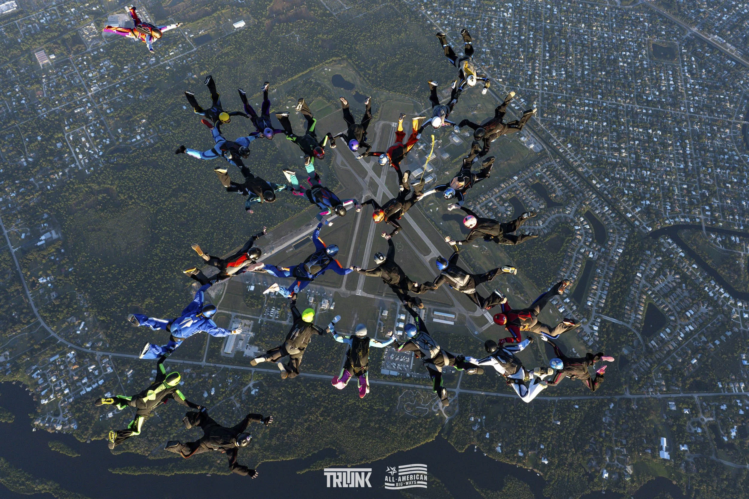Group of skydivers in colorful suits and helmets forming a star shape above a city with a runway, lakes, and roads.