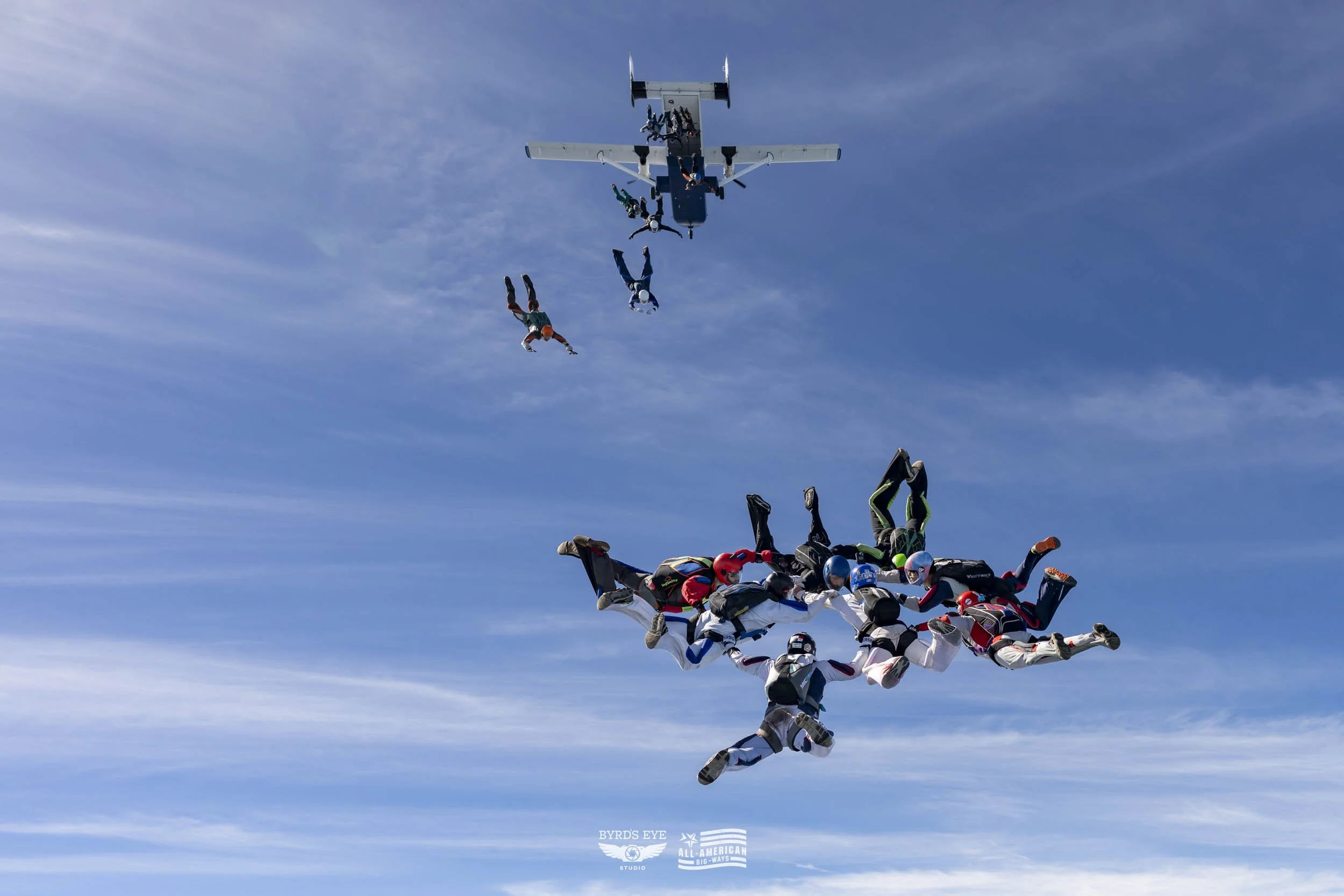 Group of skydivers in freefall, forming a cluster, with a plane flying above against a blue sky with scattered clouds.