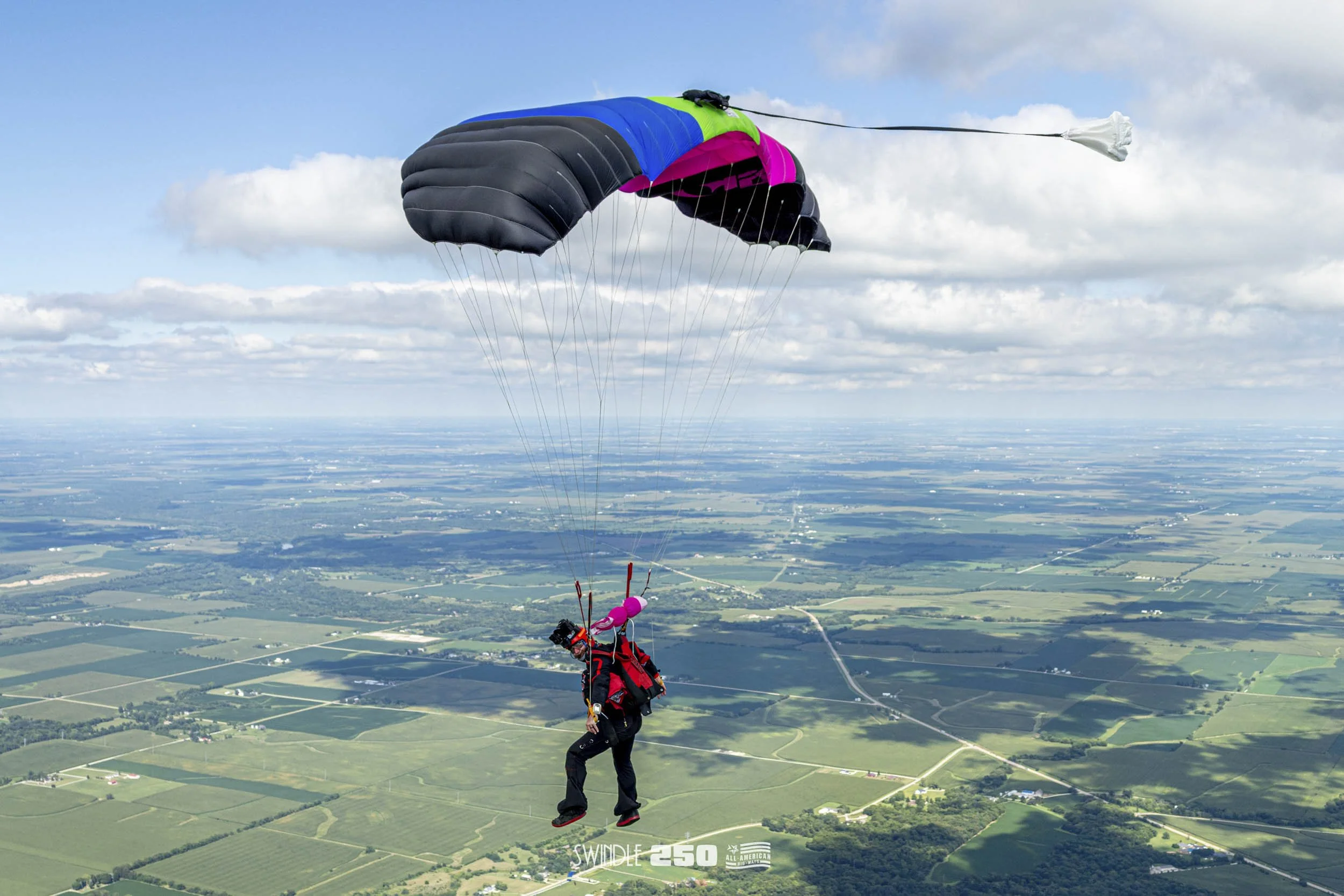 A person parachuting from a plane with a colorful parachute over a green landscape with fields and roads.