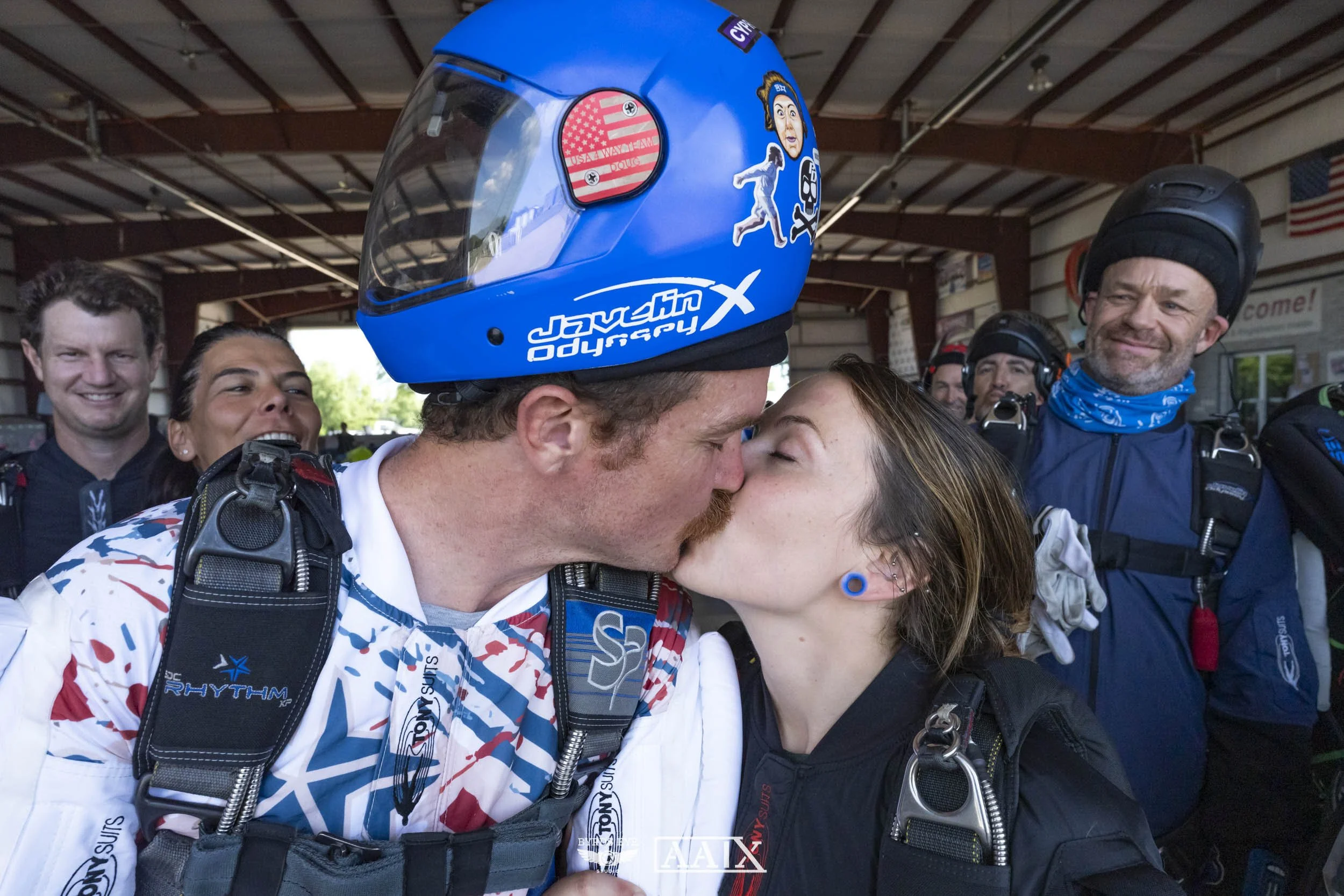 A man and woman kiss in front of a group of skydivers. The man is wearing a blue helmet and skydiving gear, and the woman has blue earrings.