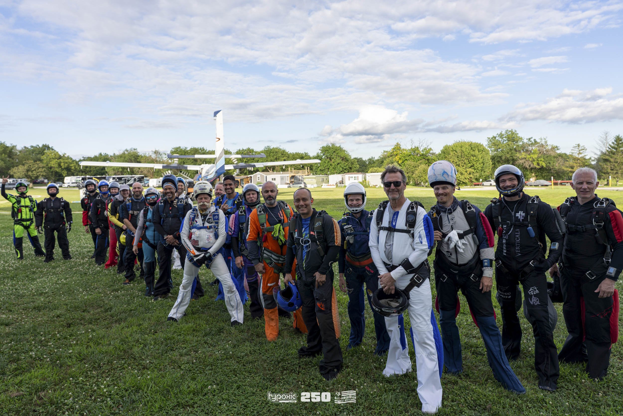 Group of skydivers in jumpsuits and helmets standing on grass field with a small airplane and sky in the background.