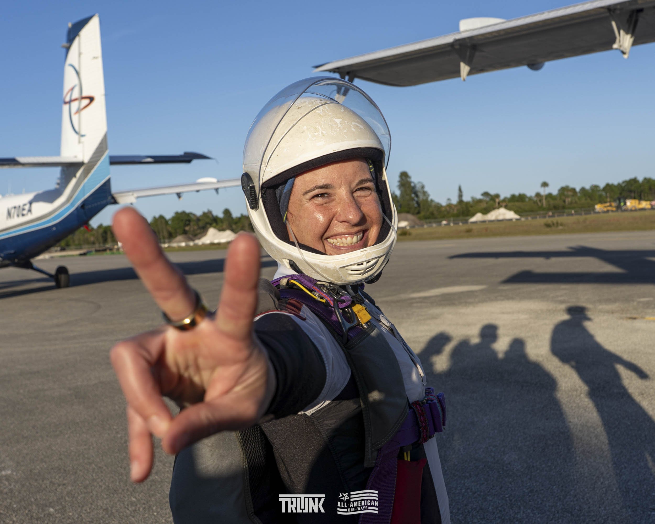 A smiling female skydiver in a helmet making a peace sign at the airport with small planes in the background.