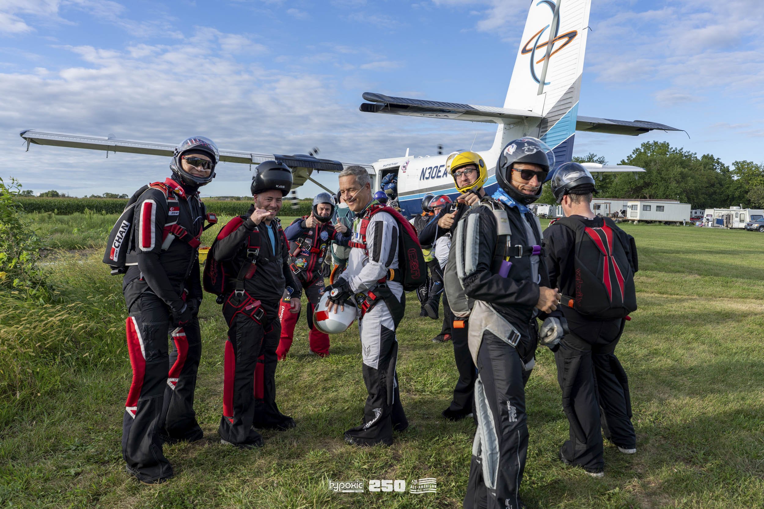 A group of skydivers in jumpsuits and helmets standing on grass near an aircraft, preparing for a jump on a clear day with a blue sky and clouds.