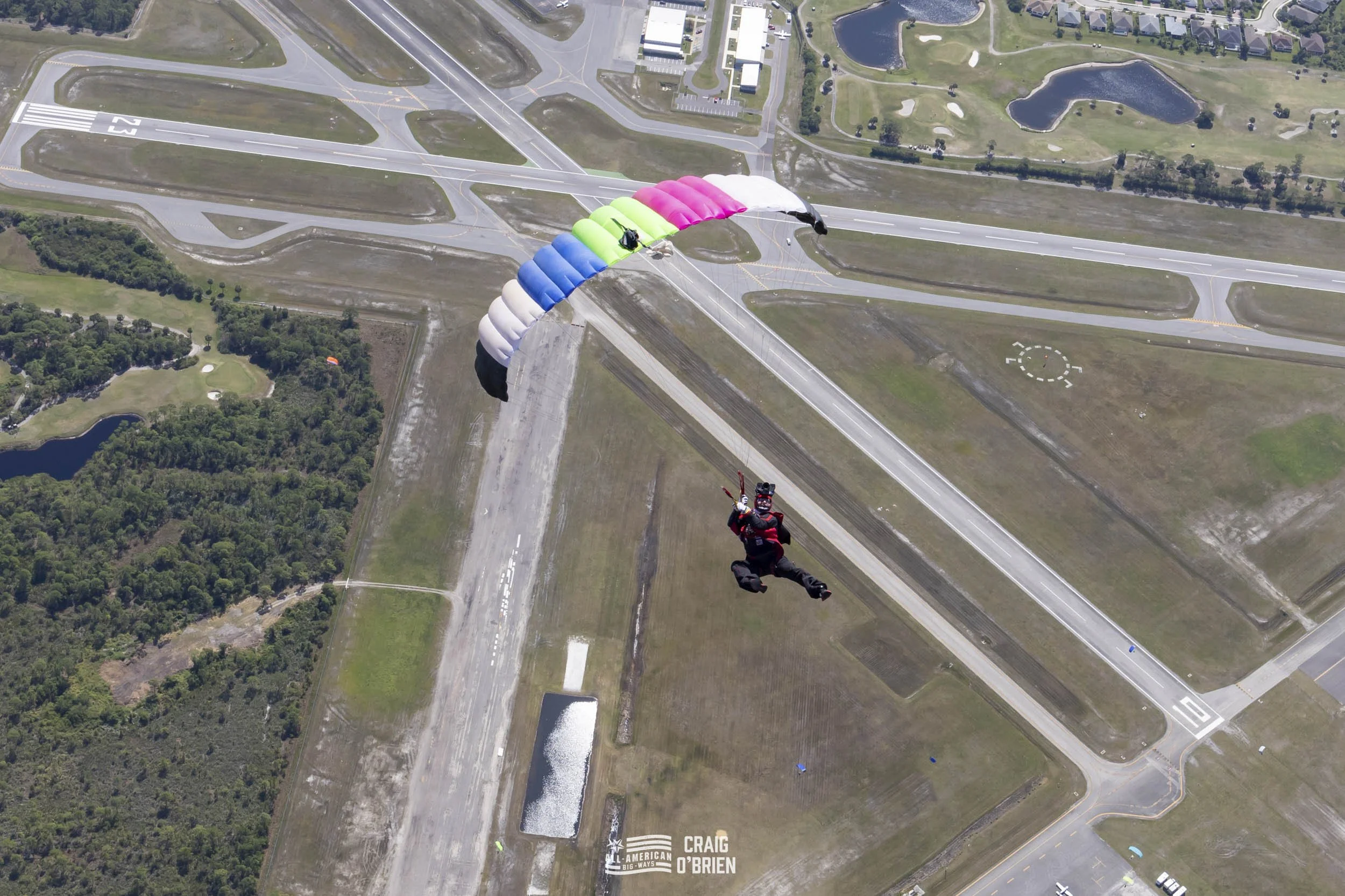 A skydiver with a colorful parachute in freefall over an airport runway, with roads, green spaces, and water bodies visible below.