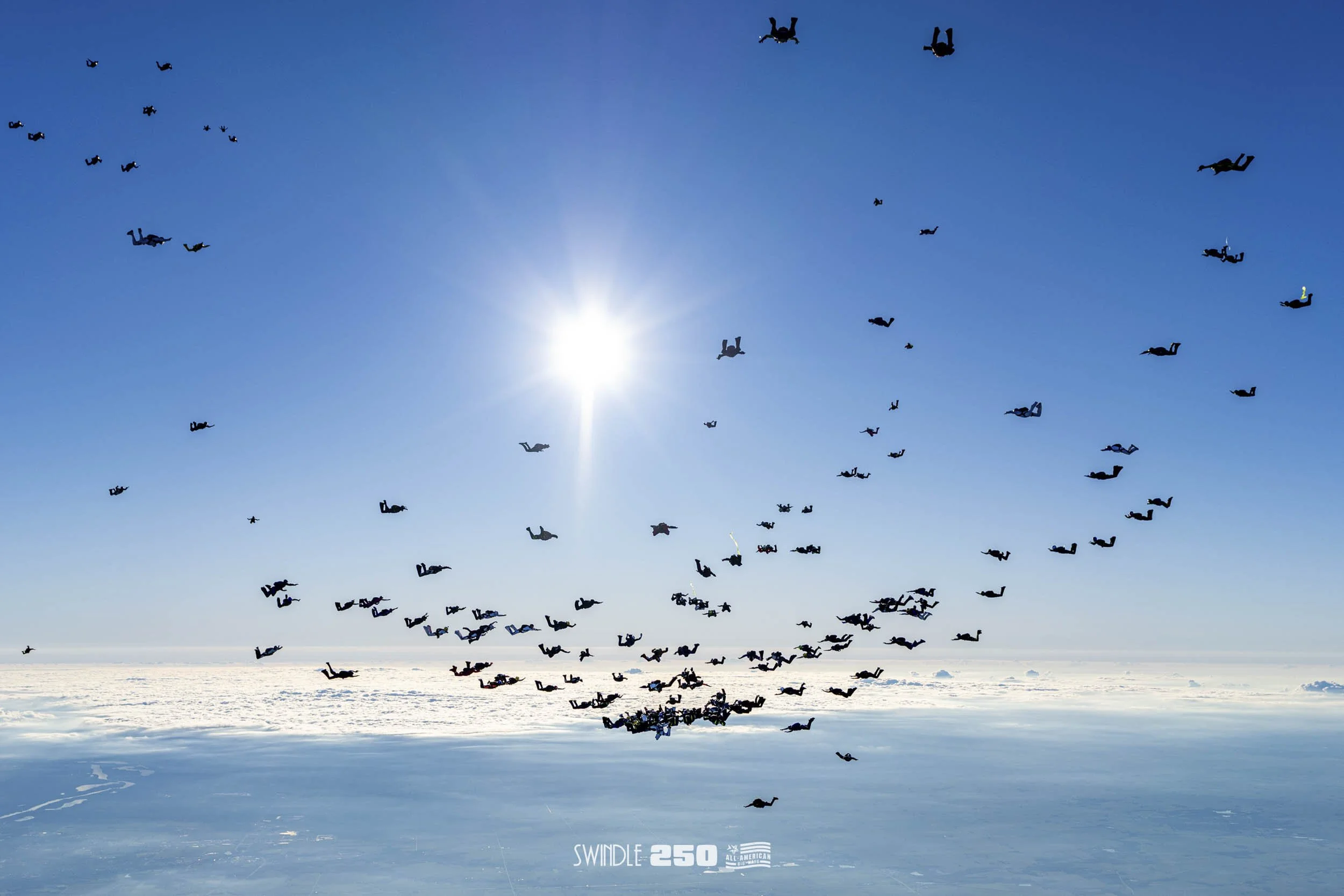 A large group of skydivers in freefall over a snow-covered landscape under a clear blue sky with the sun shining brightly.