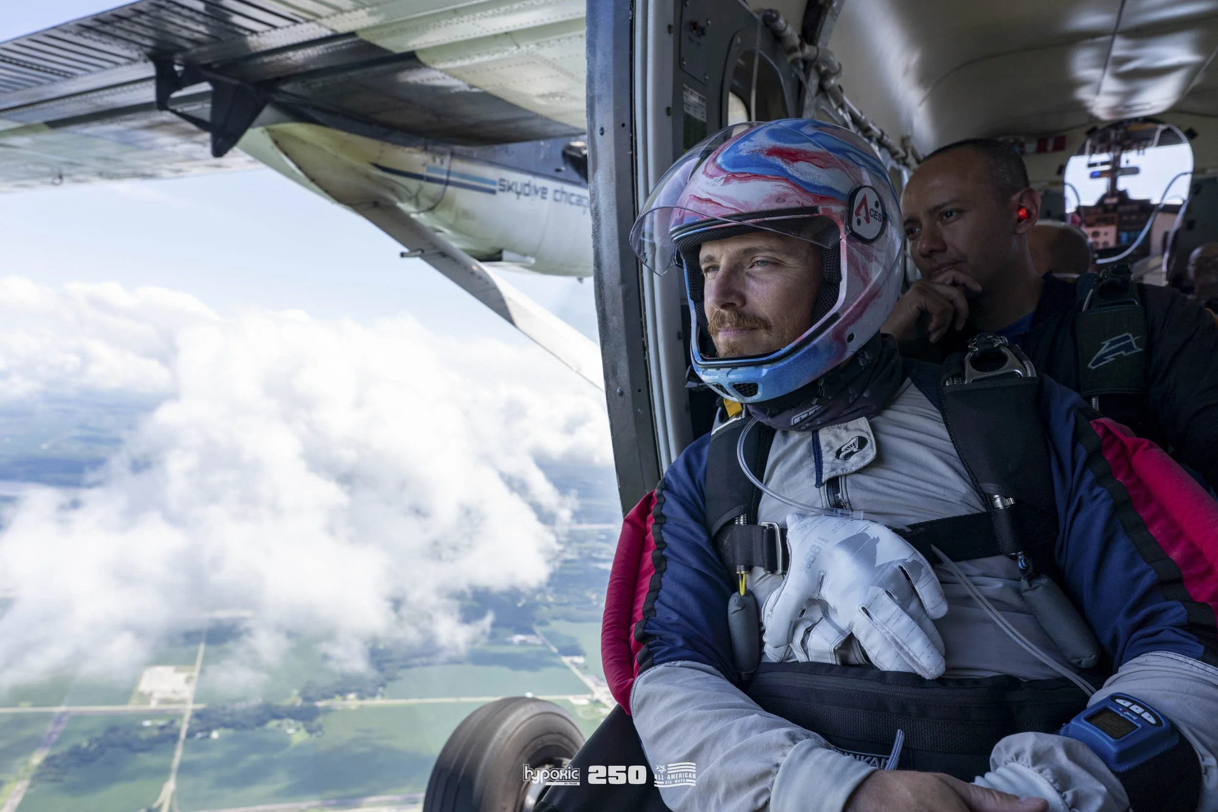 A skydiver in a jumpsuit and helmet is preparing to jump from an airplane, with cloud-filled sky and farmland below.