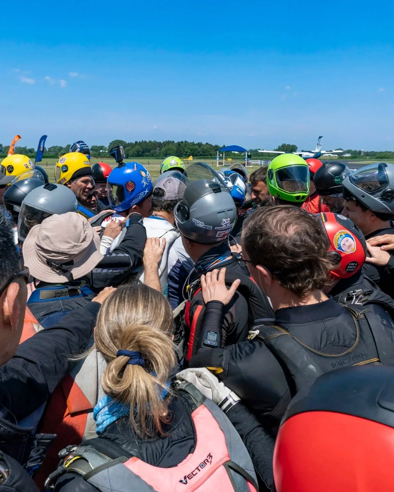 Group of skydivers in helmets huddled together on an airfield under blue skies, preparing for a jump.