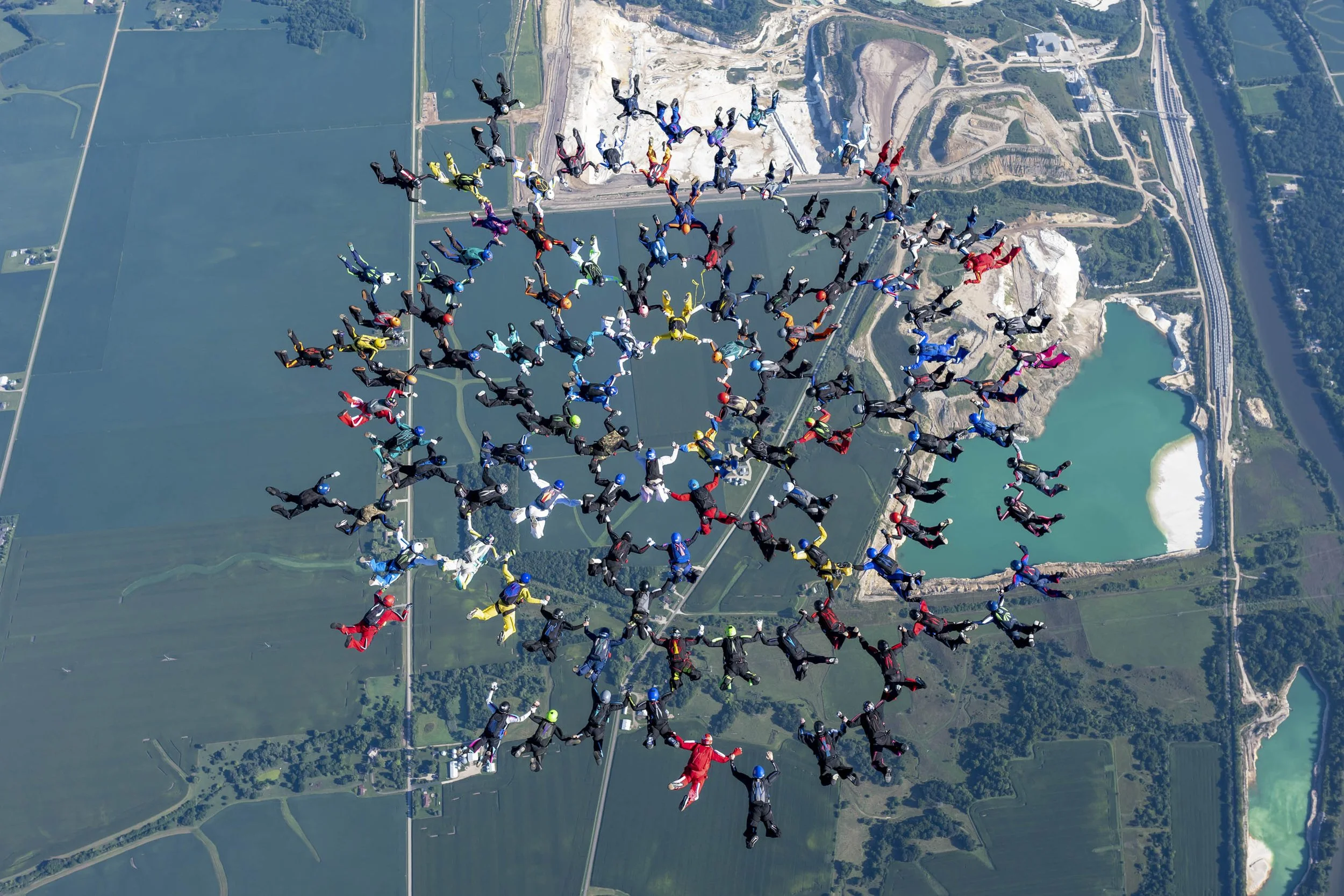A large group of skydivers in colorful jumpsuits forming a spiral pattern while mid-air over a landscape with water bodies, fields, and roads.