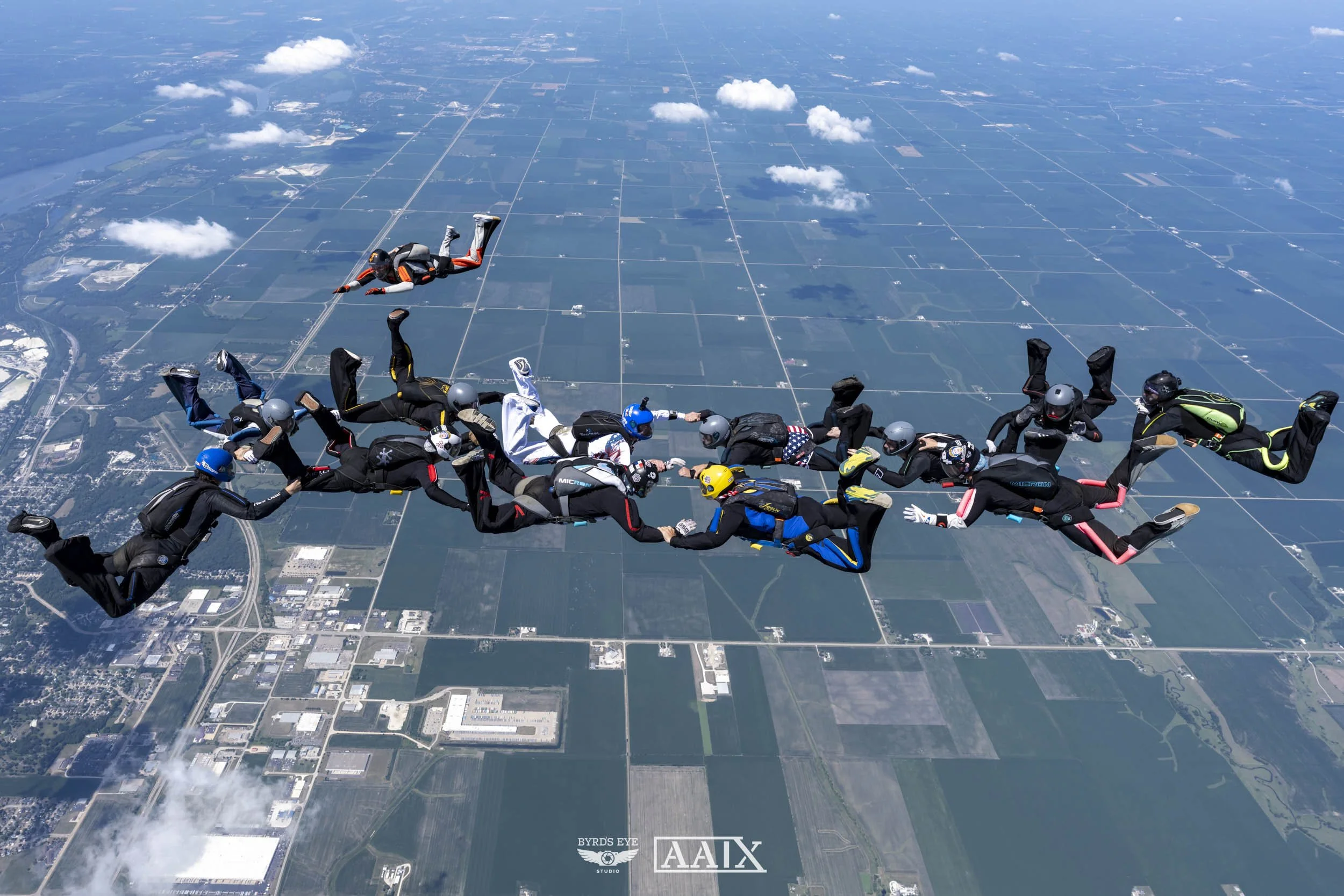 Group of skydivers holding hands in formation while free-falling over farmland and small towns.