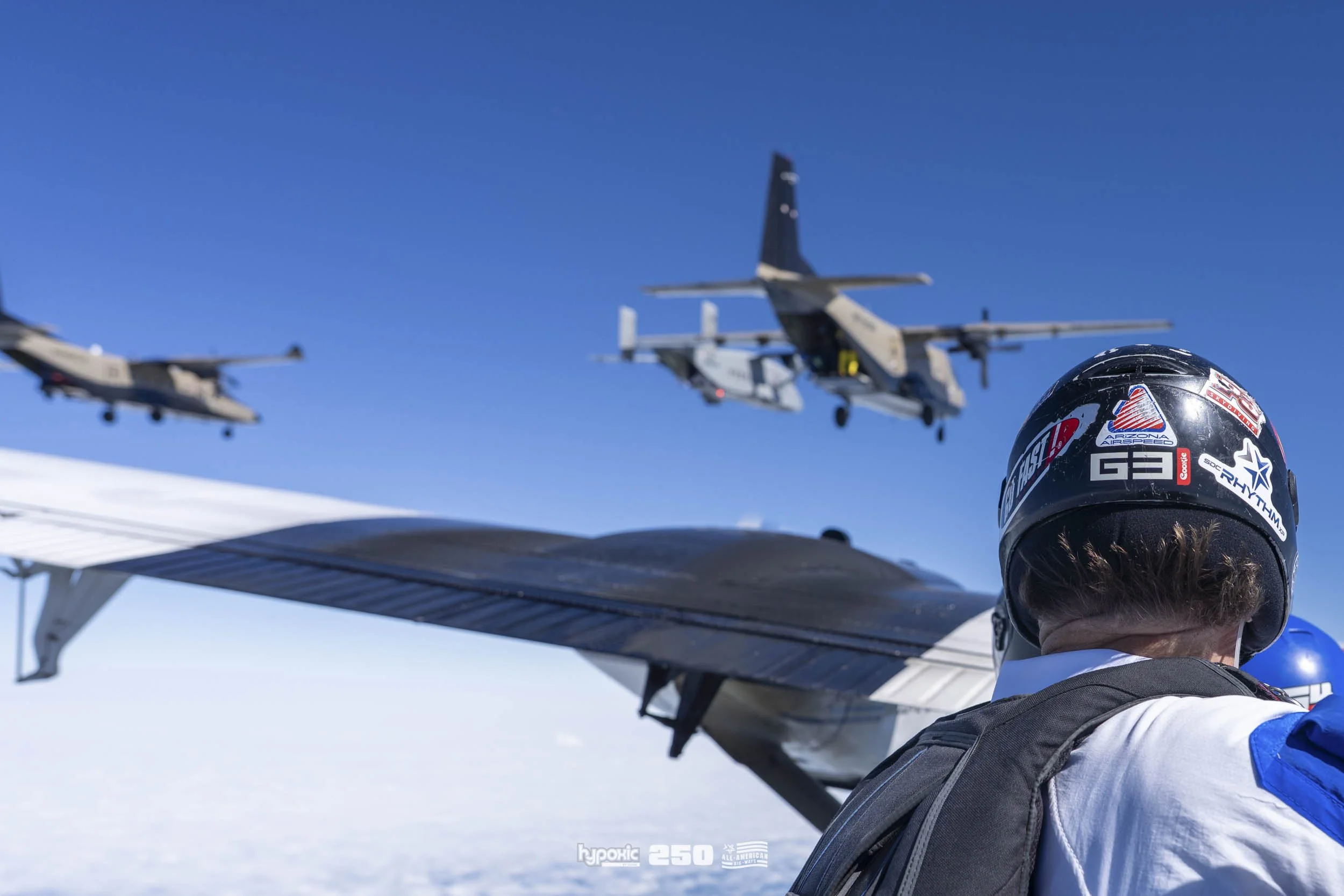A person wearing a helmet and backpack observing aircraft flying in clear blue sky above snow-covered ground.