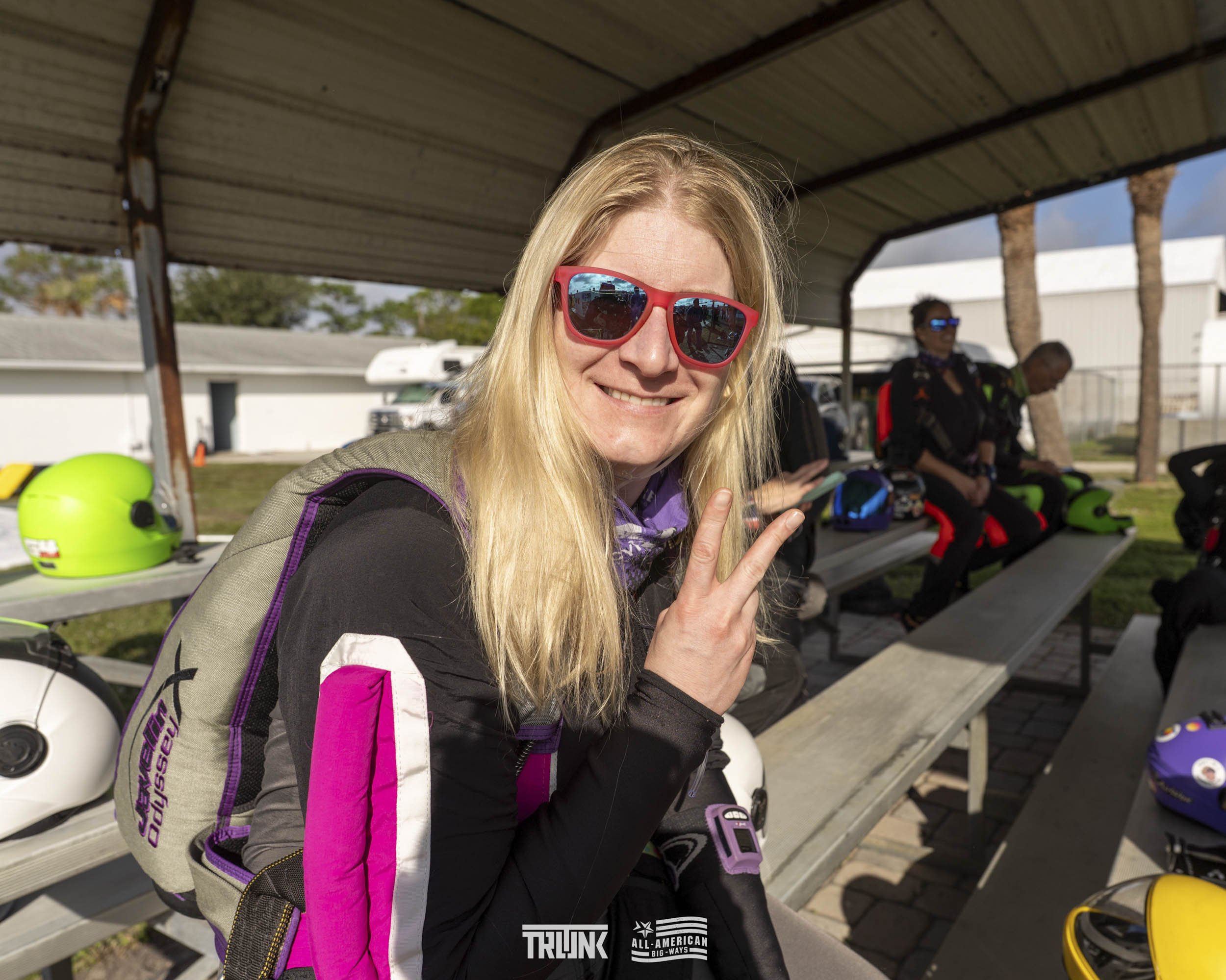 A smiling blonde woman with red sunglasses making a peace sign at a motorcycle event, with other participants and motorcycle helmets in the background.