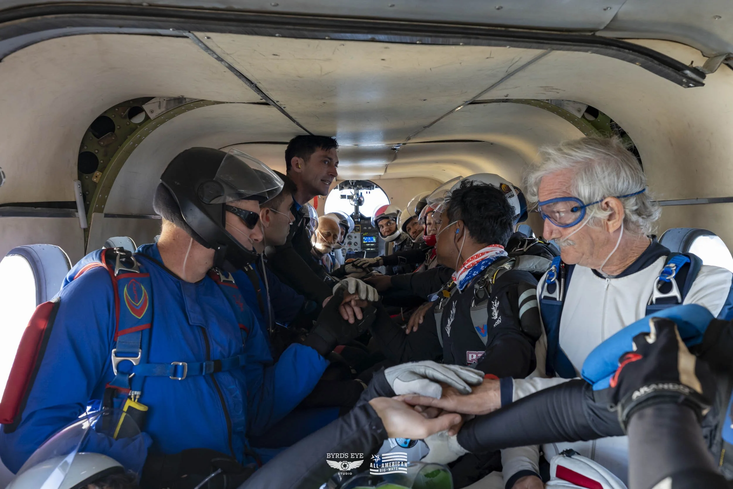 Inside a small aircraft, a group of skydivers wearing jumpsuits and helmets are sitting close together. An instructor is standing, handing out instructions or preparing the skydivers for their jump. Some are wearing goggles and ear protection. The co