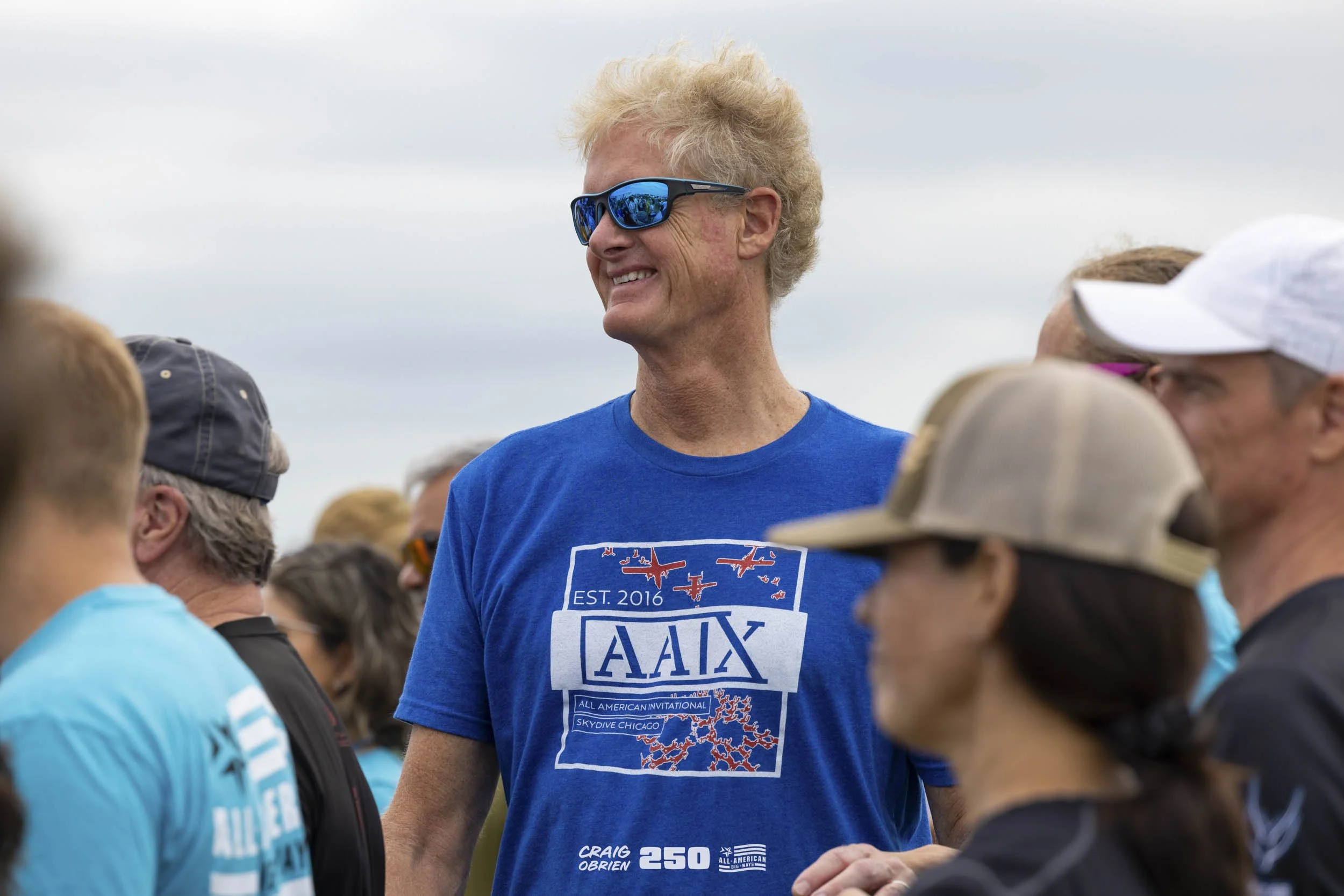A man with blonde curly hair wearing sunglasses and a blue t-shirt with the logo 'AAX' and the text 'All American Invitational Skydiving Chicago' is smiling and standing among a crowd of people outdoors under cloudy skies.