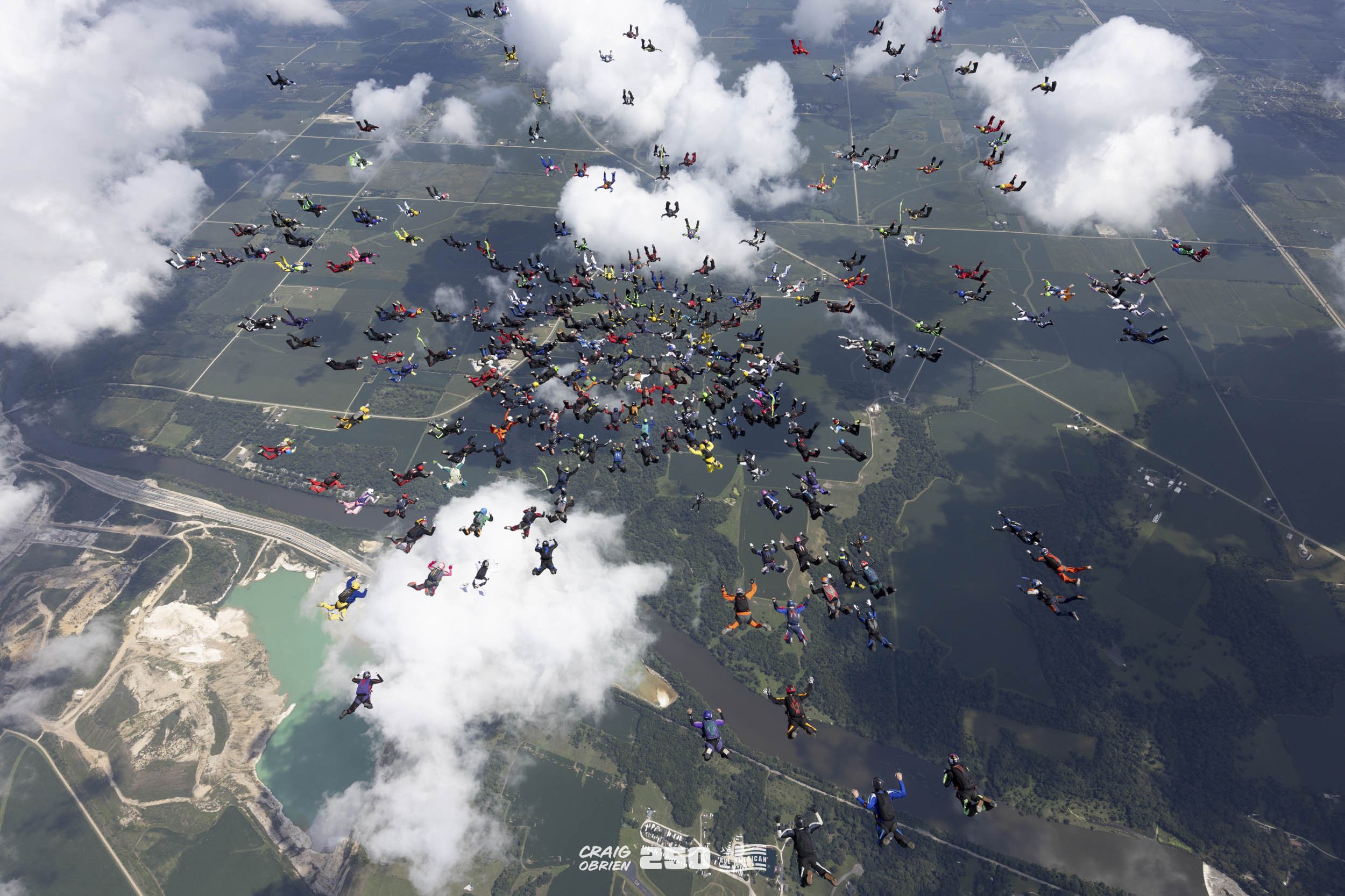 Numerous skydivers in colorful jumpsuits free-falling above a landscape with a water reservoir, clouds, and farmland.