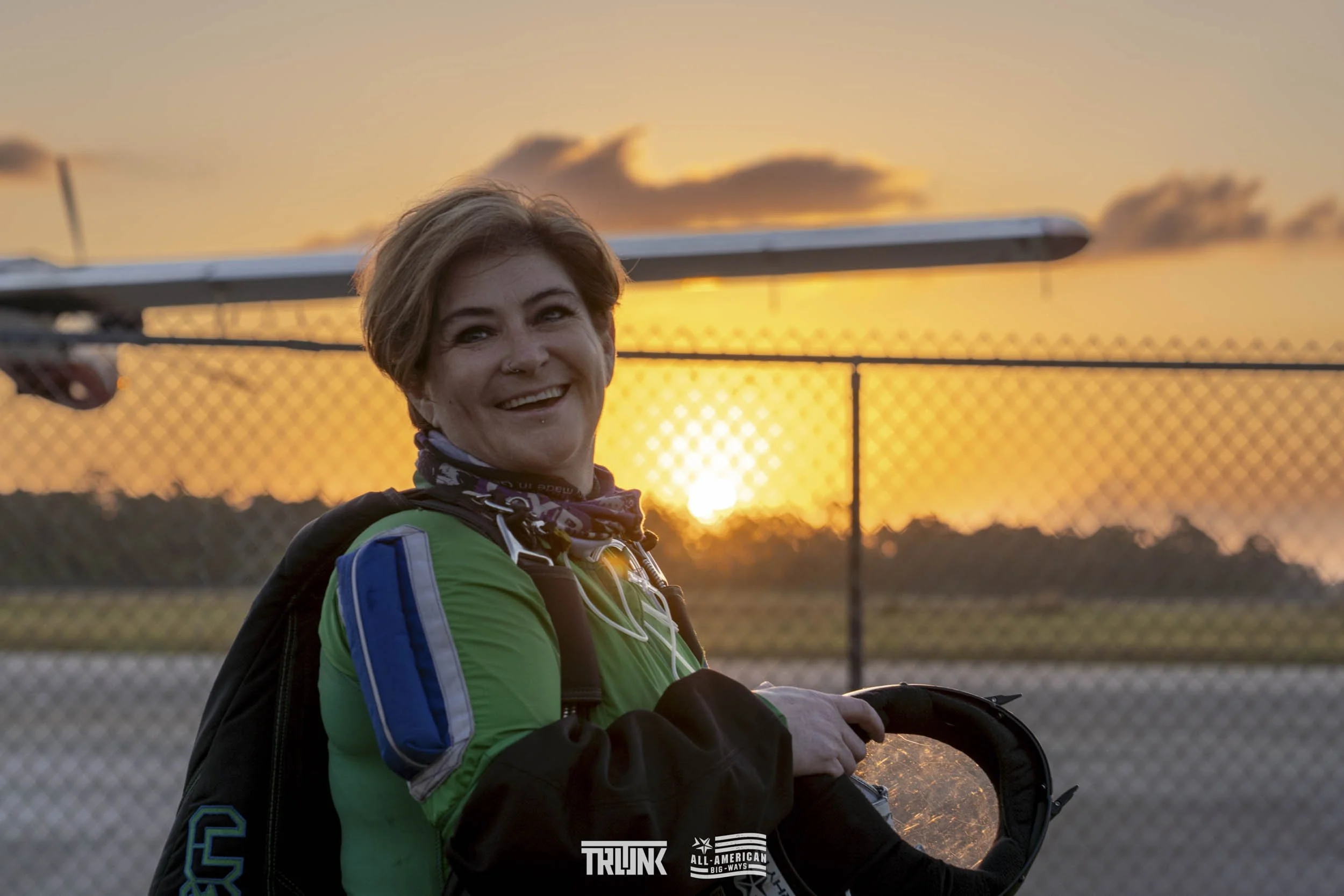 A woman in motorcycle gear smiling at sunset, holding her helmet, with a chain-link fence and trees in the background.