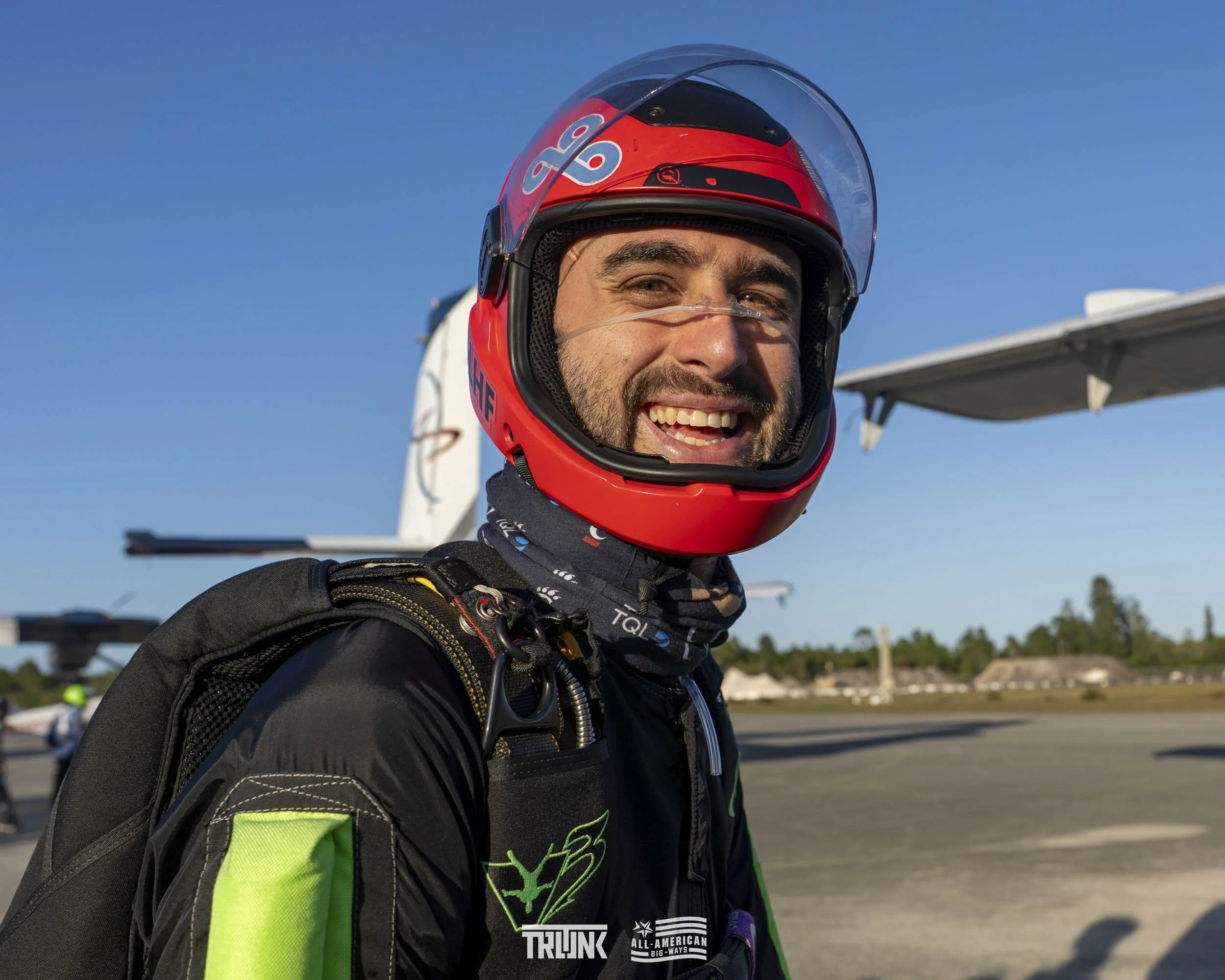 A smiling male skydiver in a black and neon green jumpsuit with a red helmet, standing on an airfield with a small airplane in the background on a clear, sunny day.
