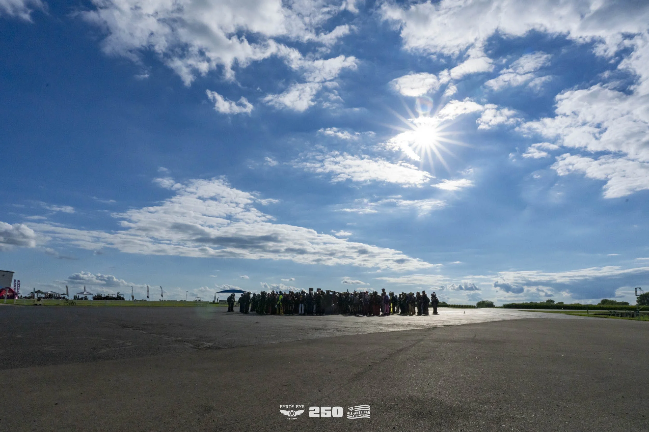 A large group of people gathered on a tarmac under a partly cloudy sky with the sun shining brightly.