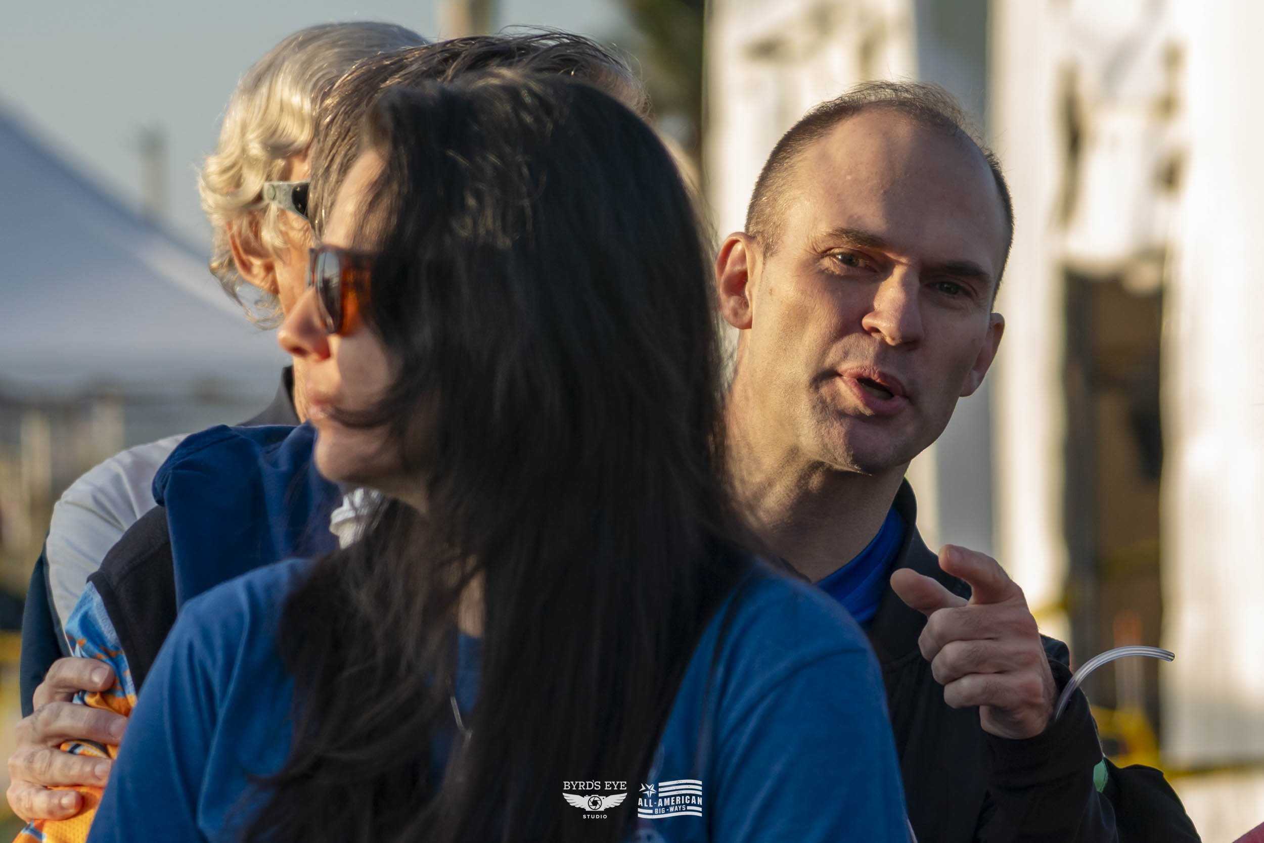 Two men and a woman engaged in conversation outdoors during daytime. The man on the right is bald, speaking, with a slight smile. The woman in front has long dark hair, and another man in the background has gray hair and sunglasses. All are casually 