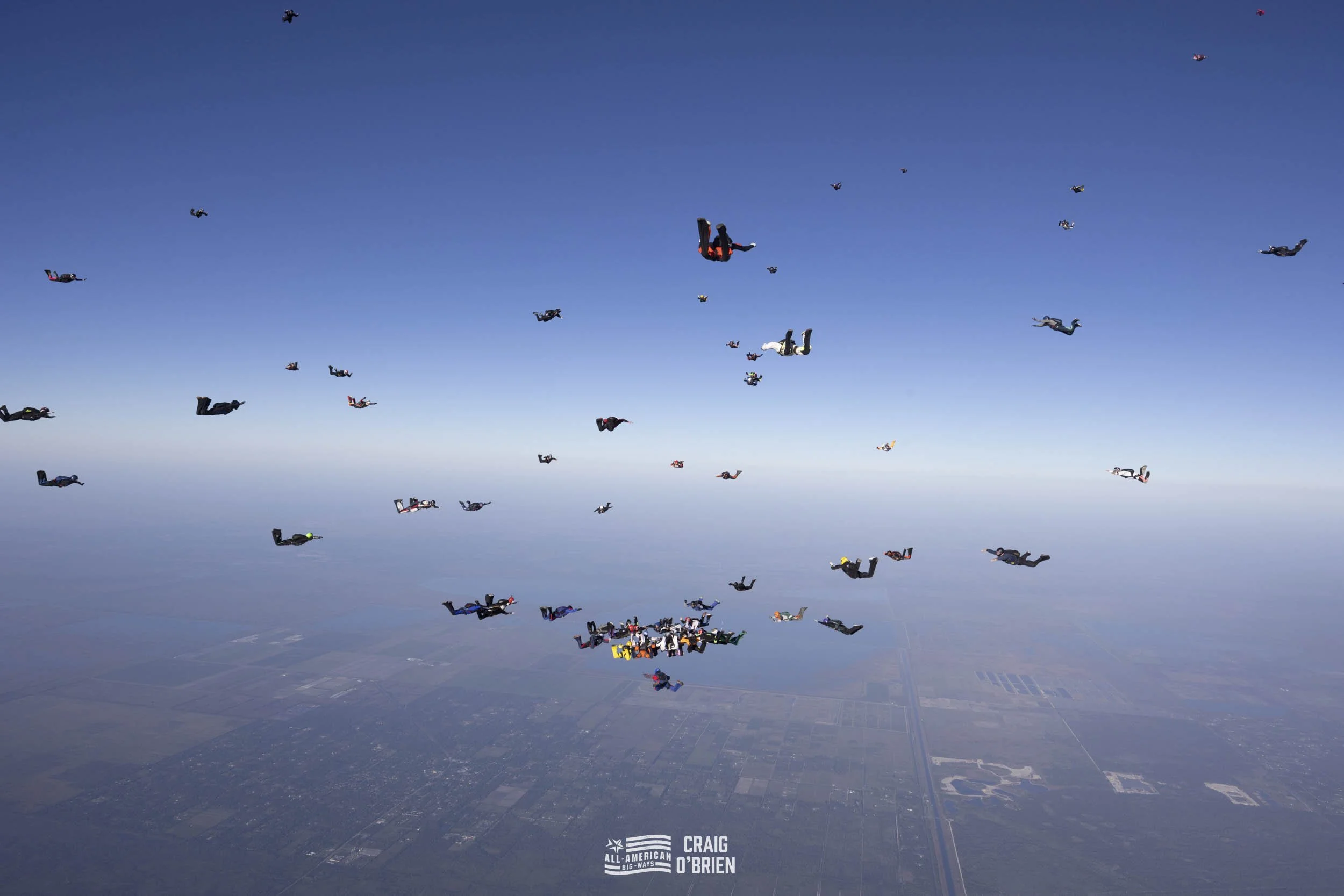 A large group of skydivers free-falling in the clear blue sky above a landscape with farmland and roads below, with the logo 'All-American Big Ways CRAIG O'BRIEN' at the bottom center.