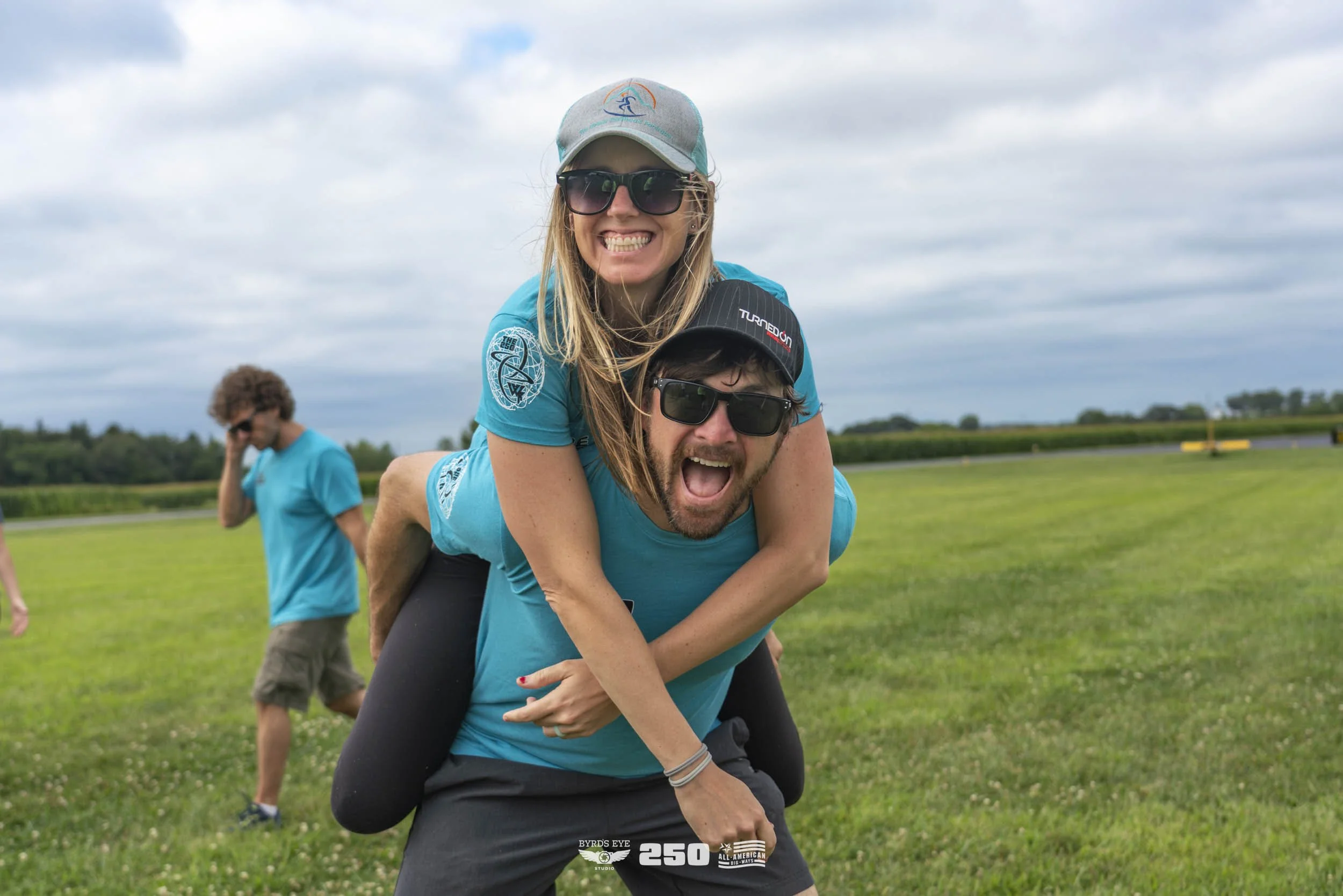 A man giving a piggyback ride to a woman on a grassy field, both smiling and wearing sunglasses and teal shirts, with other people in the background.
