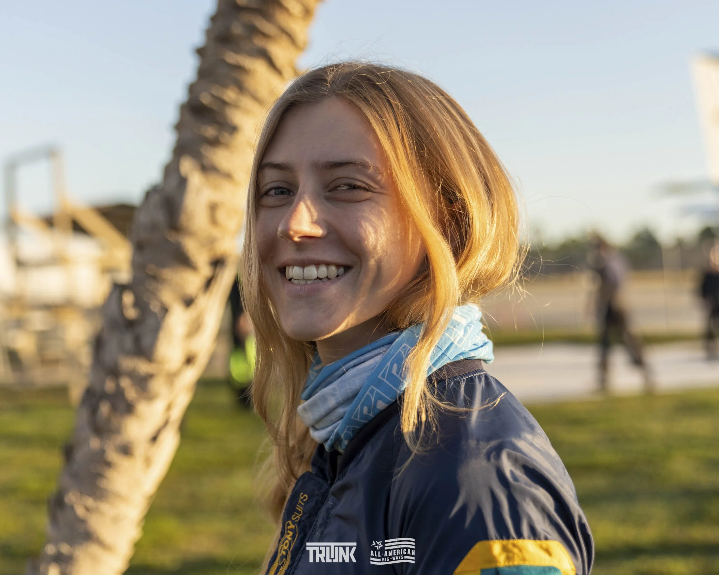 Portrait of a smiling woman with blonde hair, wearing a dark jacket and a blue neck gaiter, outdoors near a palm tree during golden hour, with a blurred background of people and an airplane in the distance.