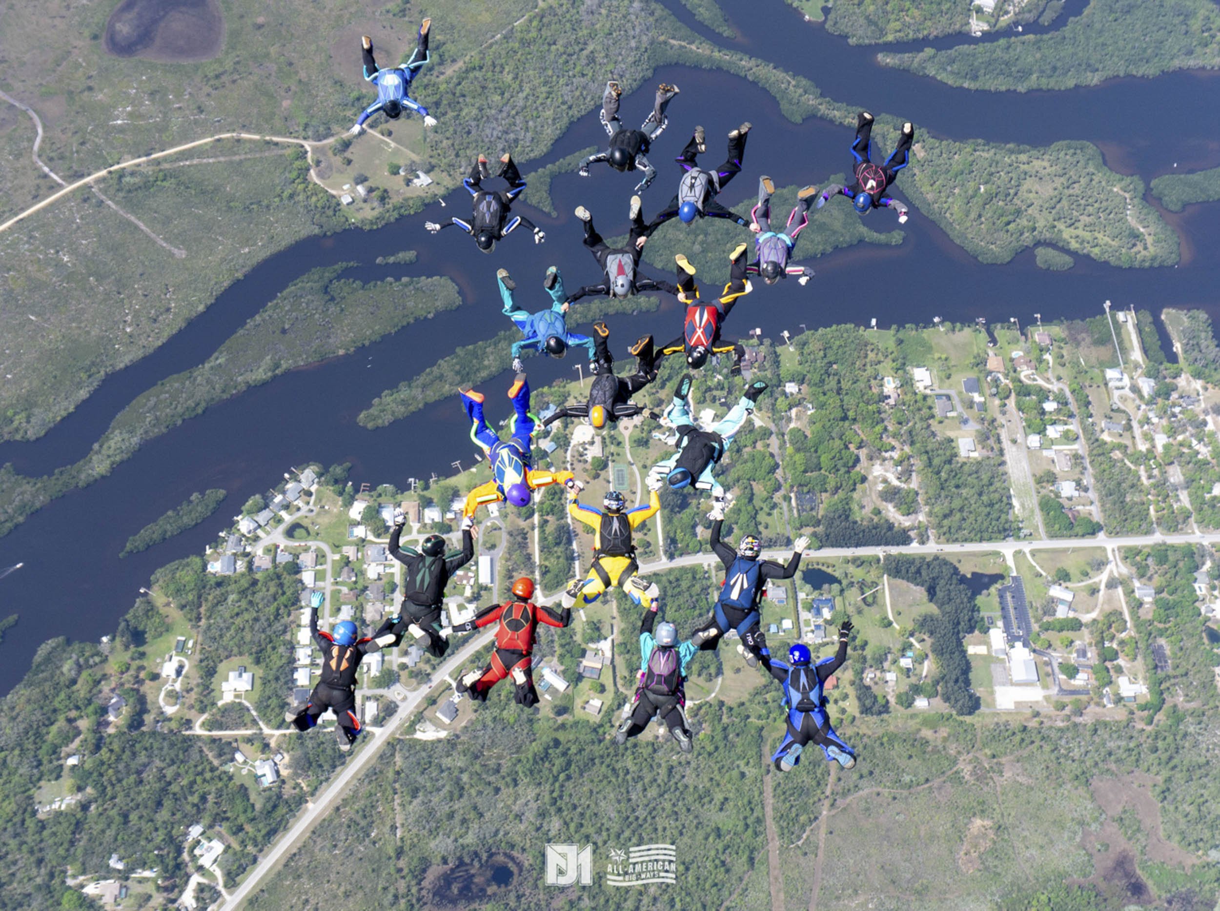 Group of skydivers in free fall over a landscape with lakes, forests, fields, and houses.