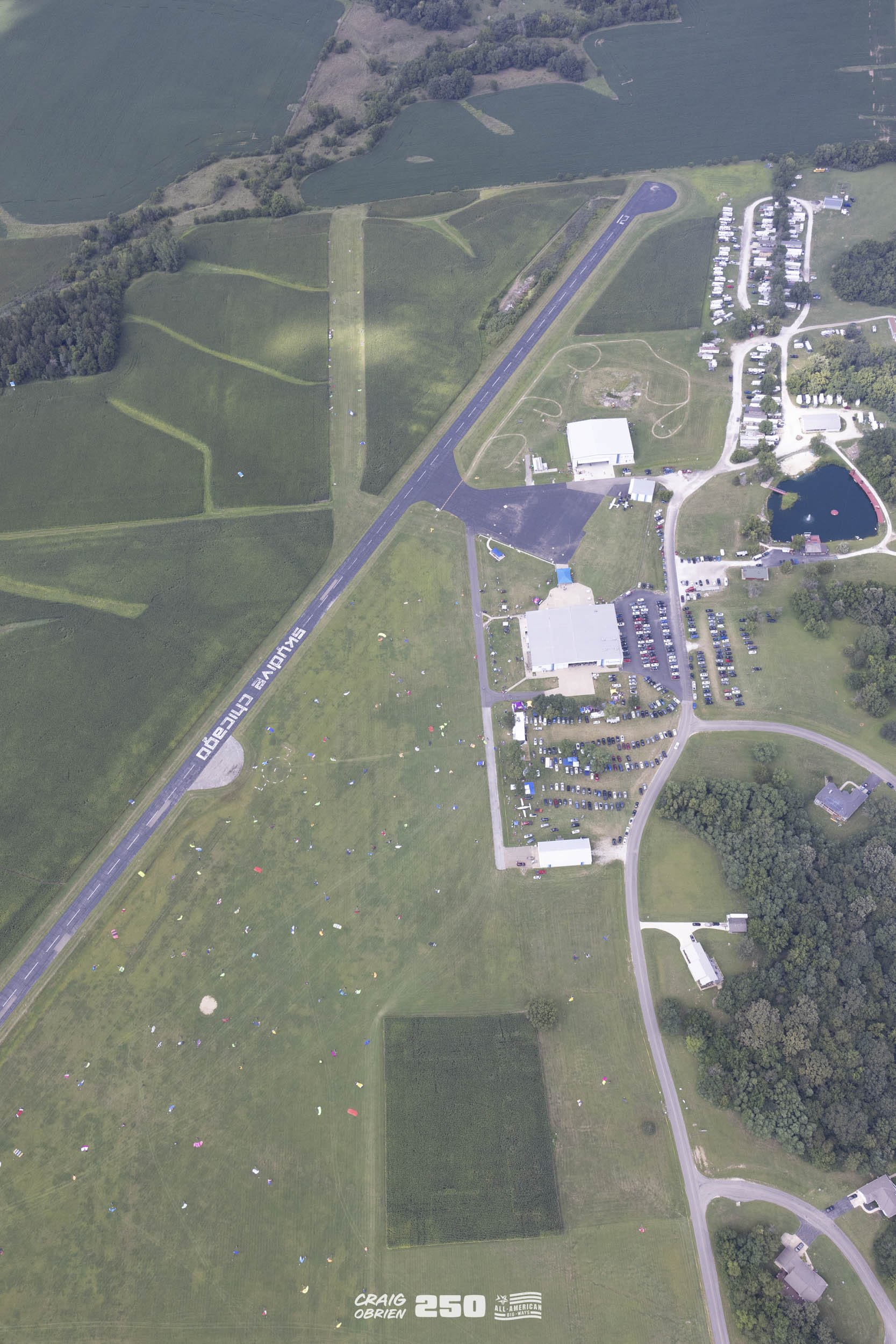 Aerial view of an airfield with a runway, parking lot, buildings, and a pond surrounded by trees and grass fields.