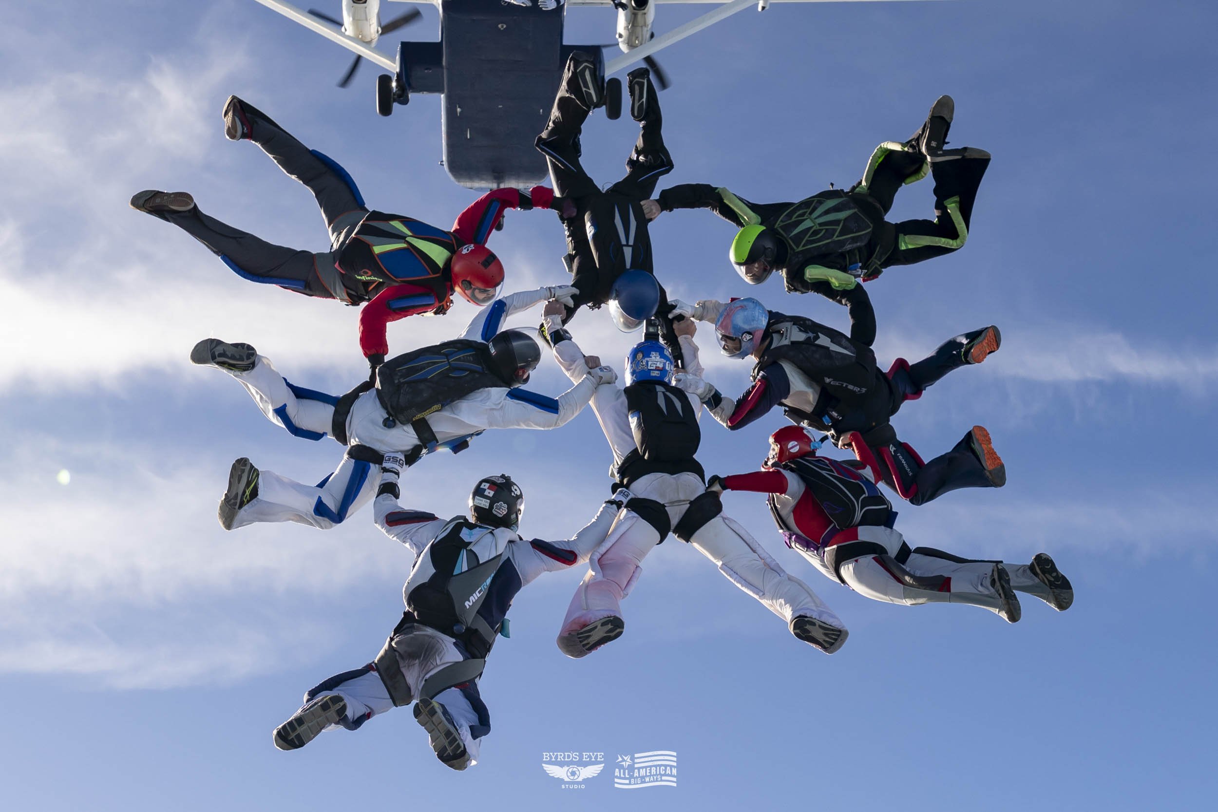Ten skydivers are holding hands in a circle during a group jump in a clear blue sky.