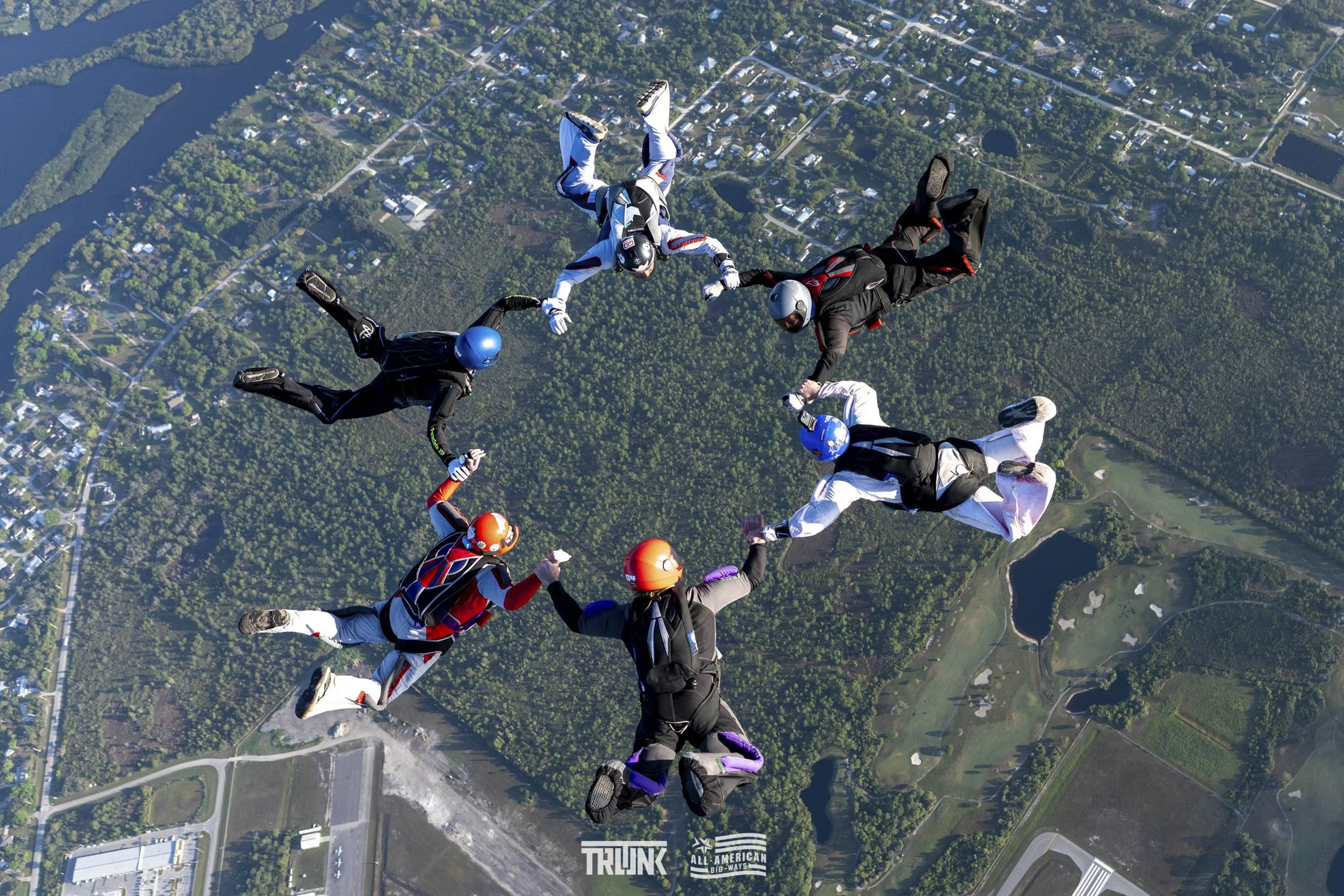 Eight skydivers in colorful jumpsuits and helmets holding hands in a circle during free fall over a green landscape with lakes and roads.
