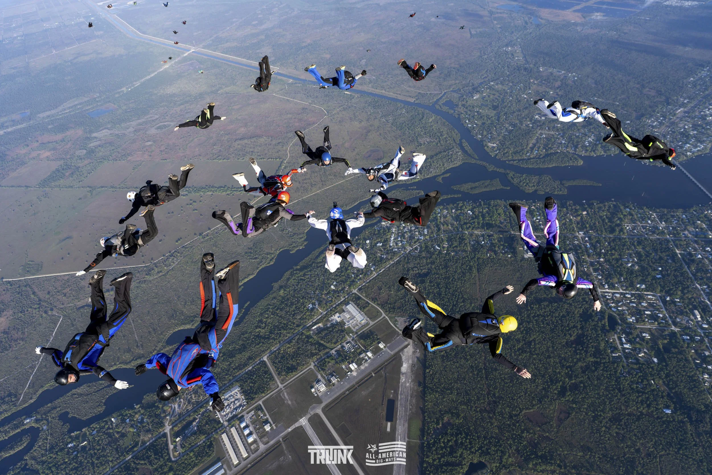 Group of skydivers free-falling in formation over a landscape with rivers, roads, and green areas