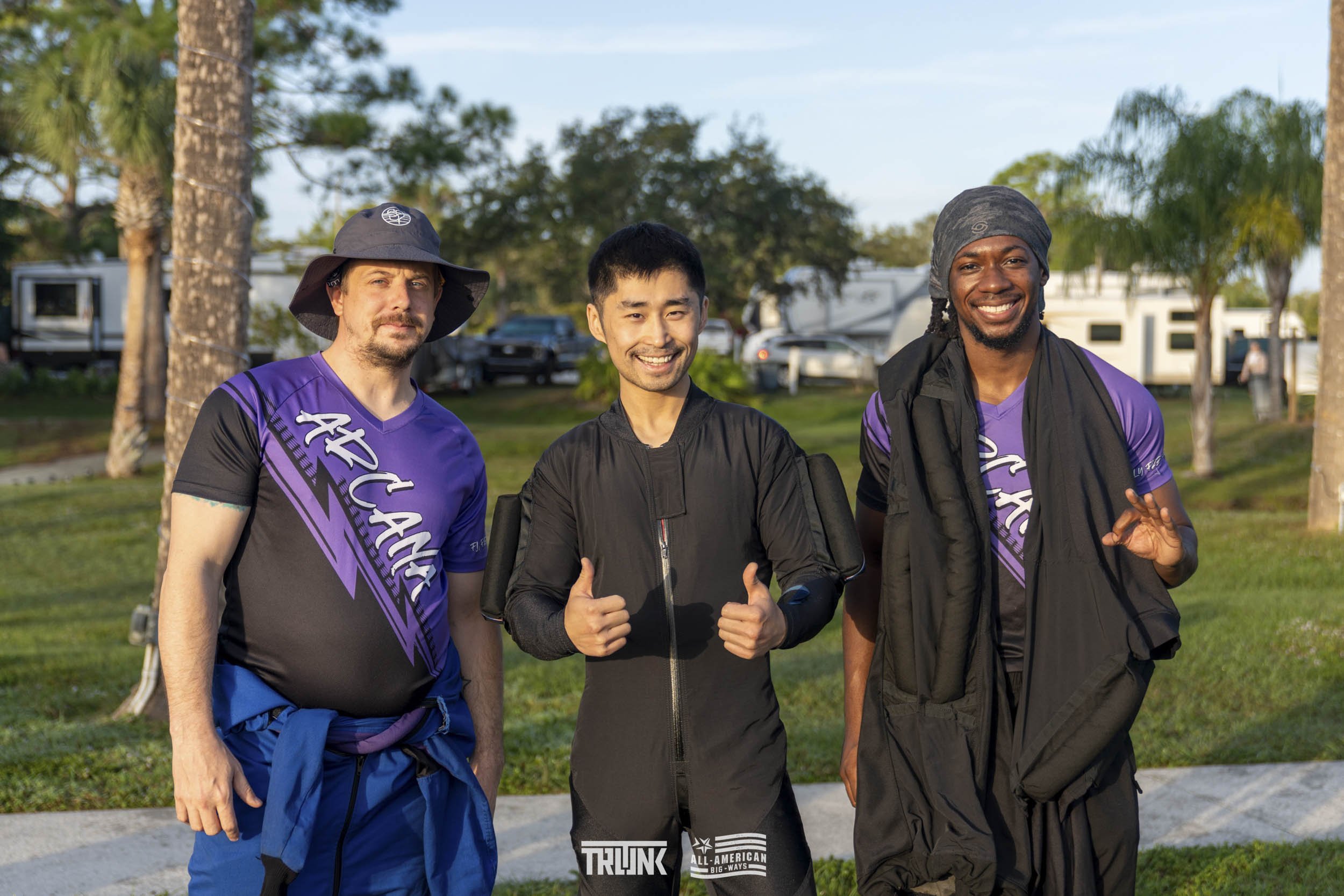 Three men standing outdoors in a park, smiling at the camera. Two are wearing purple and black athletic shirts with the word 'Attain' on them, and one in the middle is wearing a black jacket with a thumbs-up gesture. Behind them are trees, grass, and