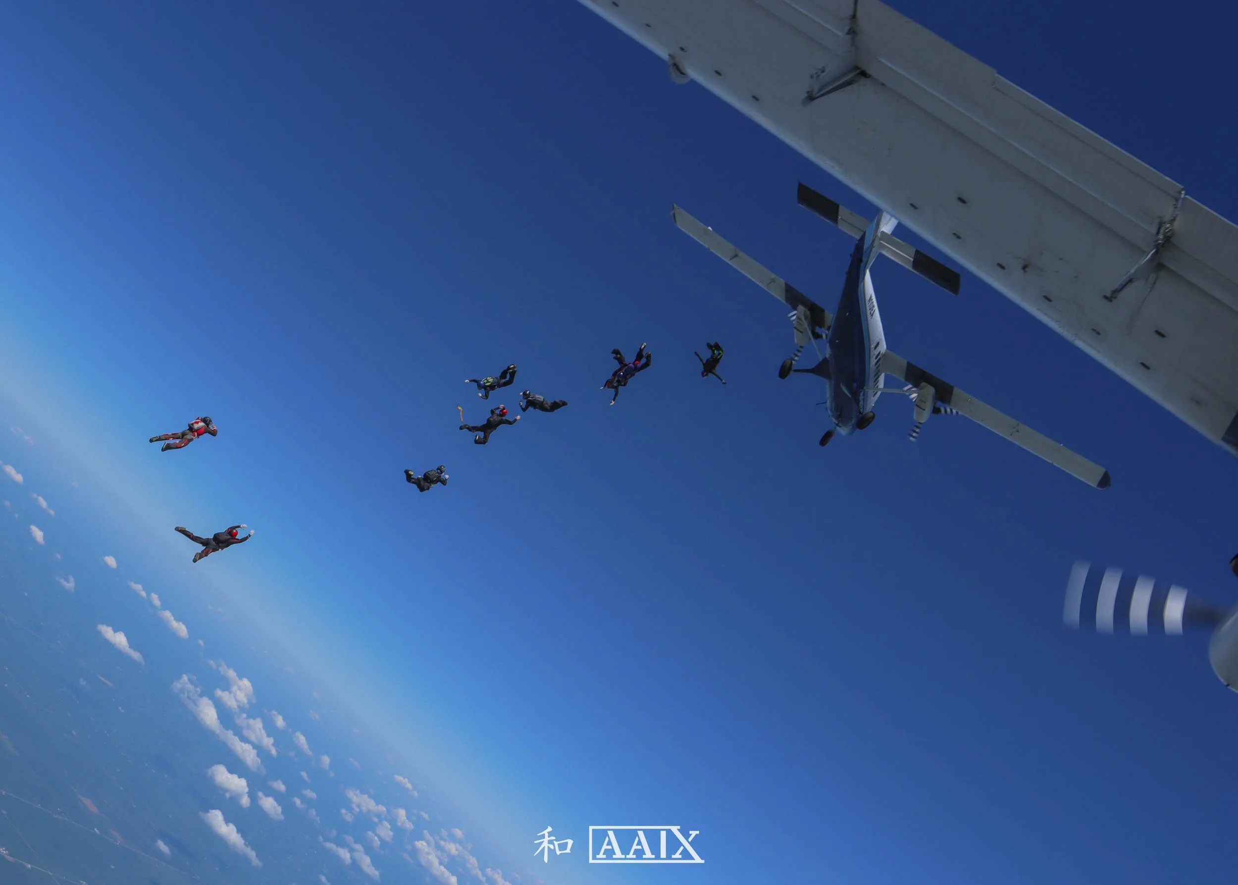 Skydivers free-fall from an airplane in a clear blue sky, with clouds below and an airplane visible near the top right corner.