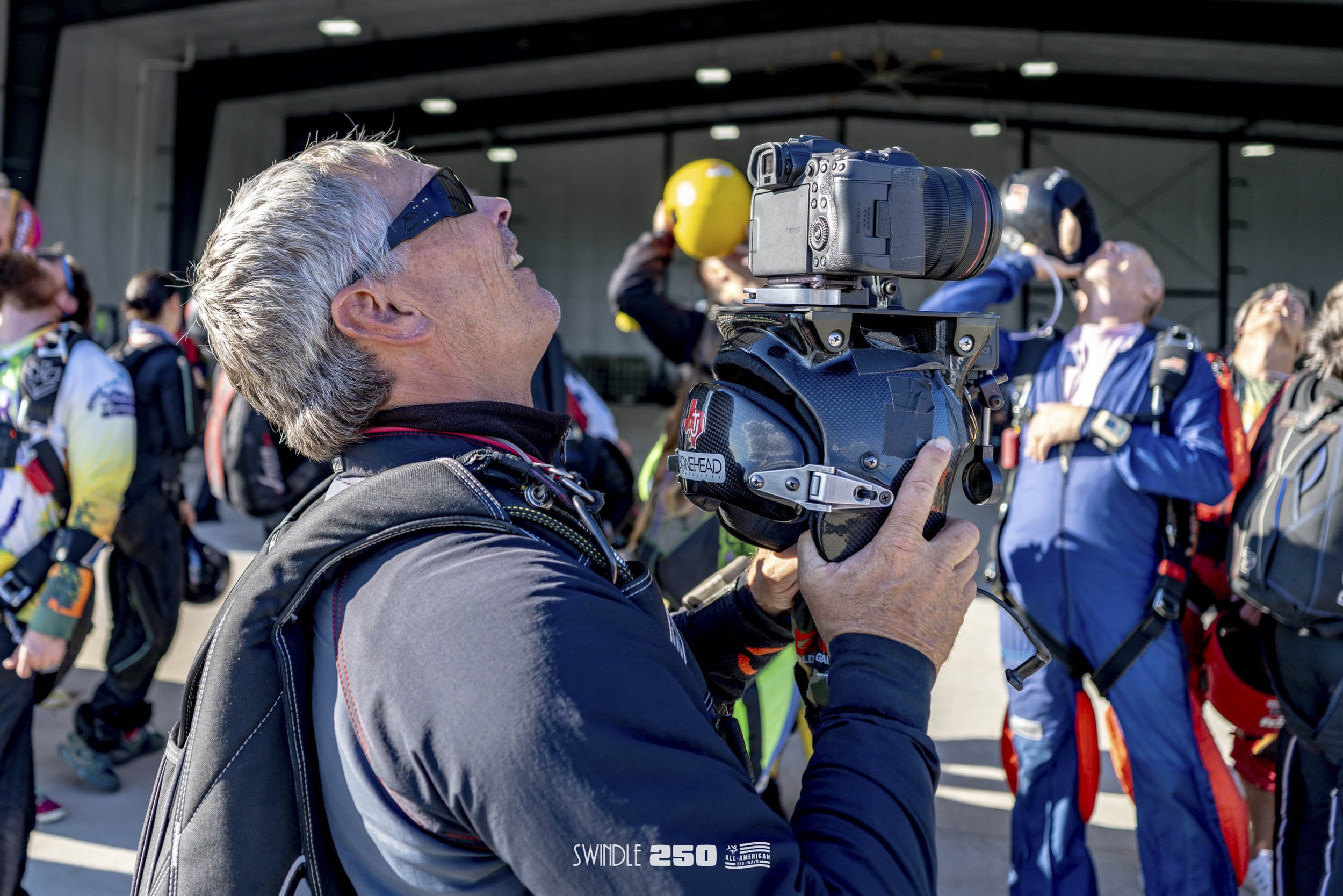 A man operating a camera at a gathering of skydivers preparing for a jump, with people in skydiving jumpsuits and helmets in the background.