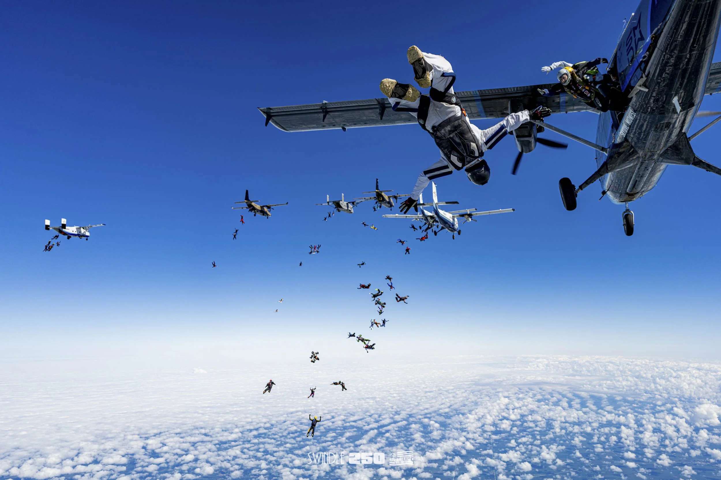 Military aircraft releasing paratroopers into the sky over a cloud-covered landscape during a jump exercise.