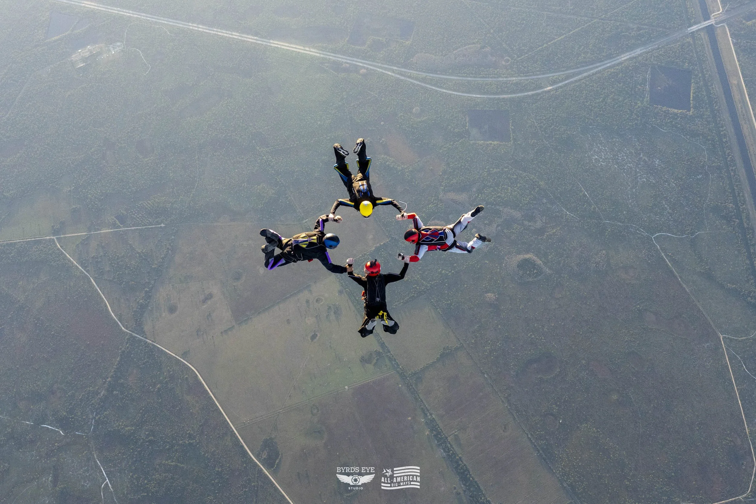Four skydivers in freefall connected in a star formation with the ground visible below.